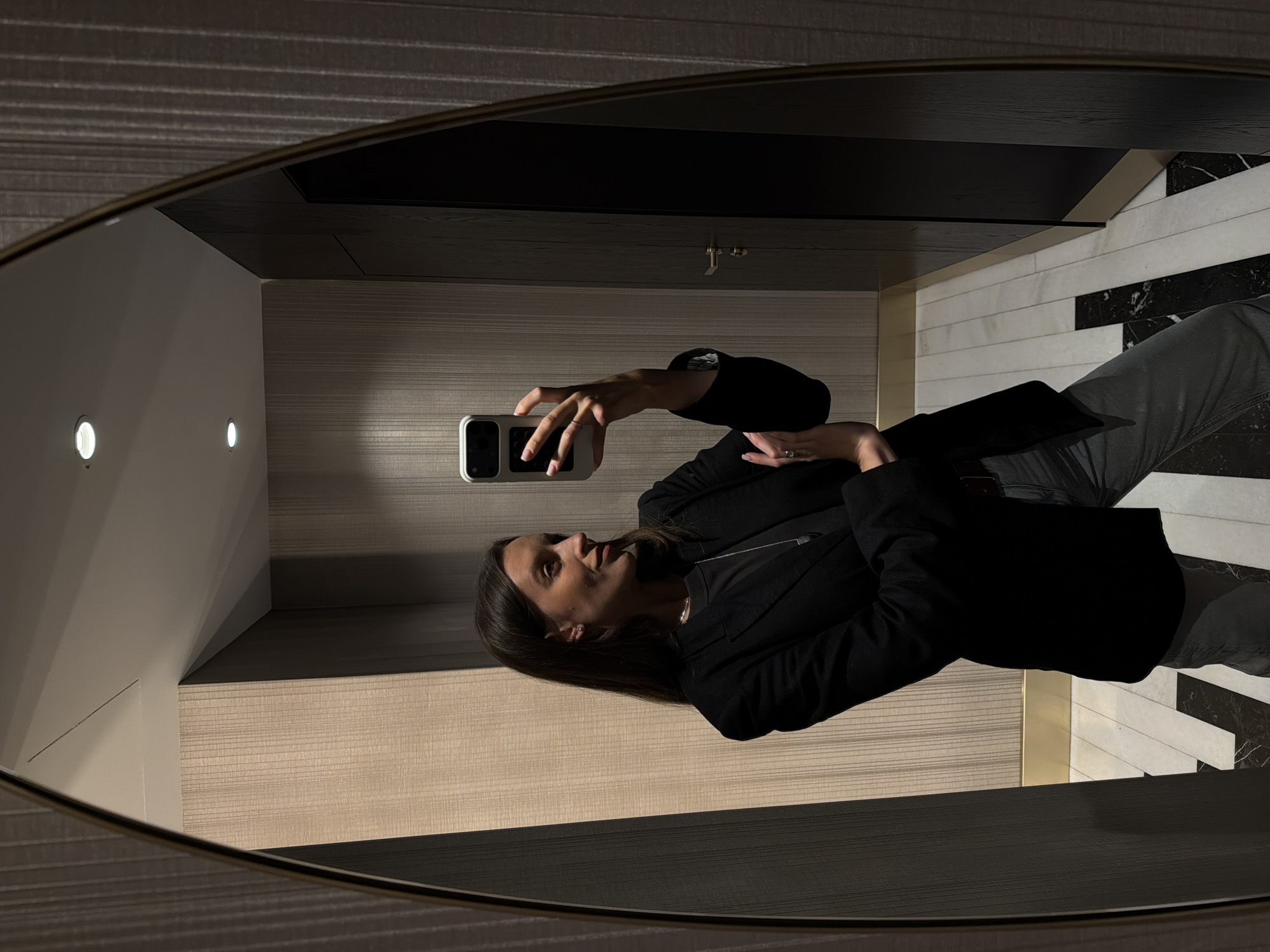 A person in a dark blazer and grey trousers takes a selfie in a mirror, likely in an interior location. The floor has a black and white marble pattern. The lighting is artificial, with two circular lights on the wall. The location is unknown, and there is no information that the image was taken in Budva, Montenegro.