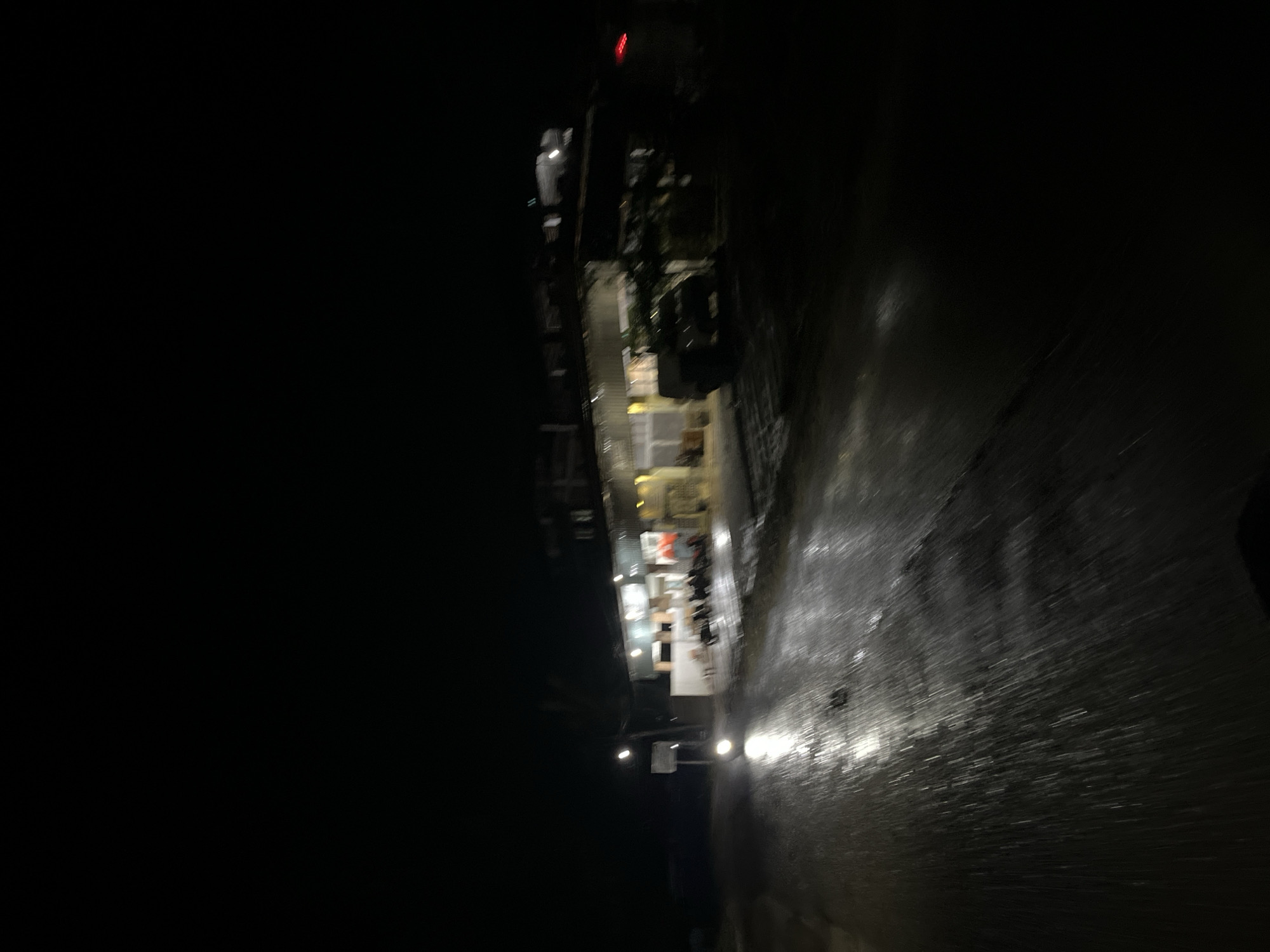 The image captures a night scene in Benuaraja, Indonesia, likely a waterfront area. The perspective is from above, looking down at an angle. The left side of the image is predominantly dark, suggesting a lack of ambient light or possibly open water.
On the right side, a lit pathway or street runs diagonally. The surface appears wet, reflecting the light from several street lamps and lights emanating from a building complex. This building complex, partially visible and illuminated, seems to have multiple floors and windows, some of which are brightly lit, revealing interior spaces that appear to be commercial or residential. One section of the building has a red sign or light visible at the top. The architecture suggests a modern commercial development.
The wet surface of the pathway indicates recent rain or that the area is consistently damp, a common condition in tropical climates. The streaks of light on the wet surface could also be from passing vehicles, though no distinct vehicles are clearly visible.
The overall mood is quiet and somewhat mysterious due to the darkness and the limited visibility of details. The illuminated buildings and pathway suggest human activity, but there are no clearly discernible people in the frame. The scene is likely taking place in a developed area within Benuaraja, possibly a harbor, marina, or a street adjacent to a body of water. The time of day is unequivocally night. There is no visible text in the image.