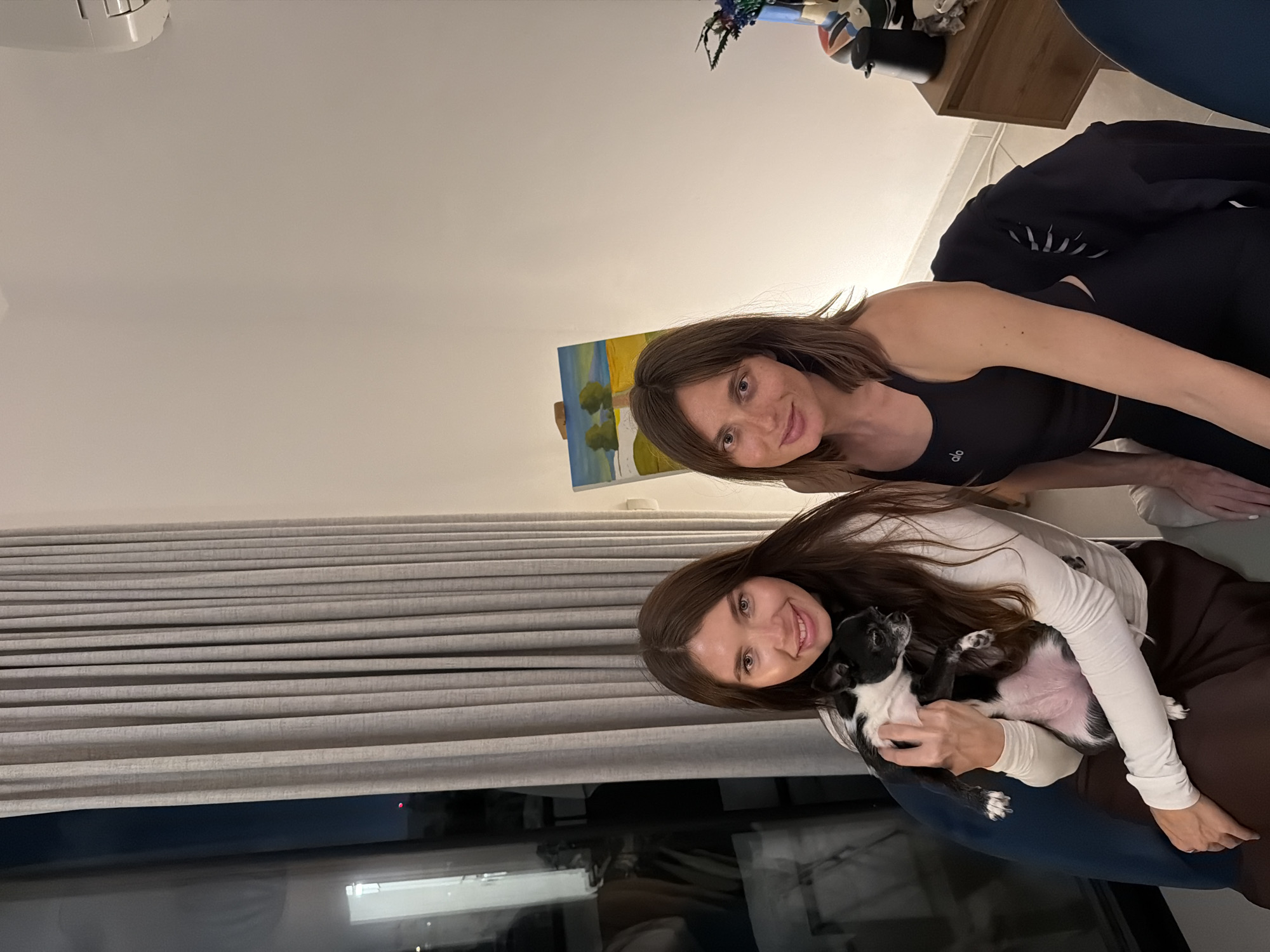 The image shows two women indoors. One woman is holding a black and white dog. The location appears to be Tel-Aviv, Israel, based on the urban nighttime view through the window. The background includes a painting on an easel and other household objects.