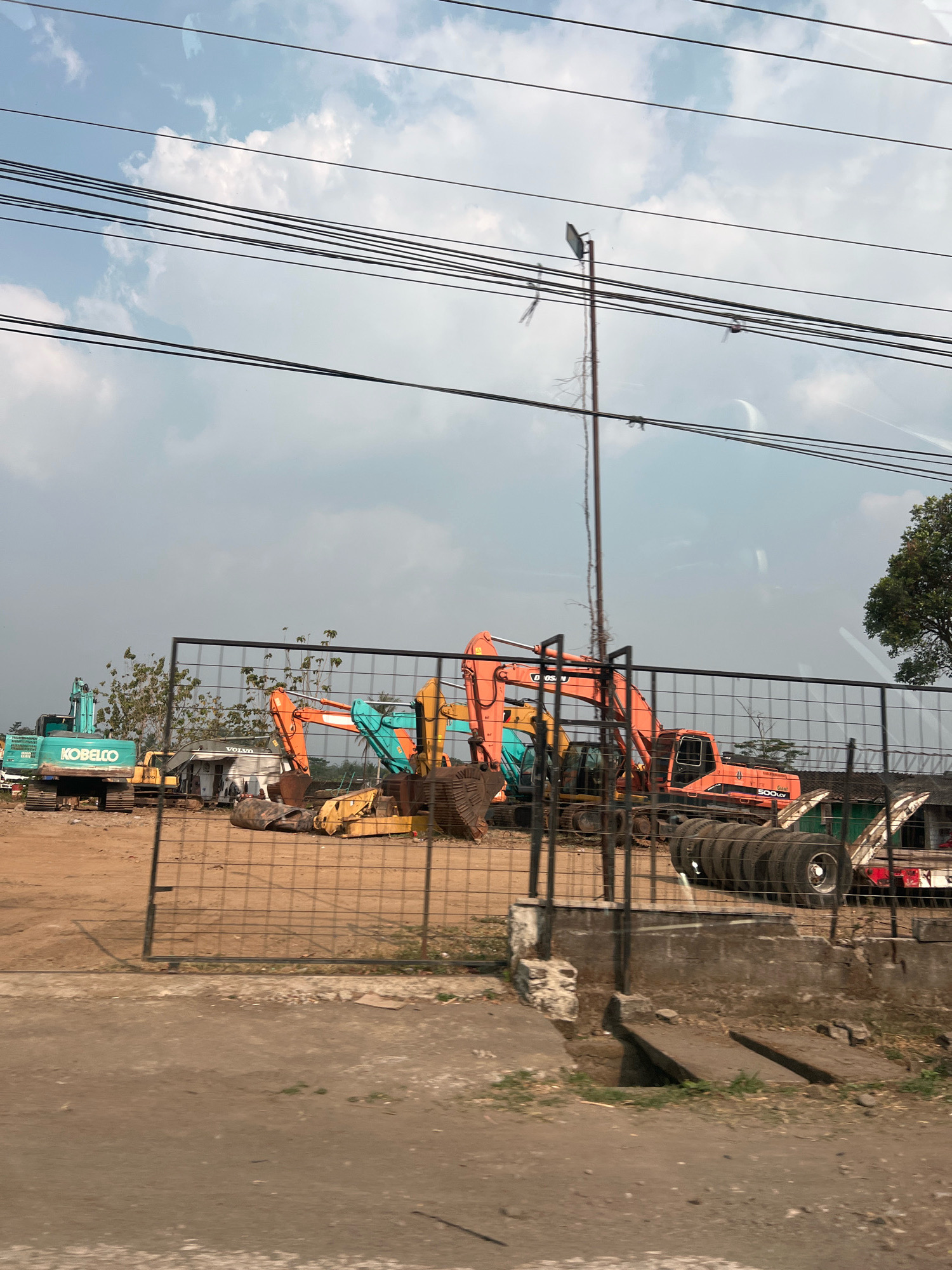 This image depicts an outdoor construction equipment storage lot in Secang, Indonesia. The scene is dominated by several large excavators, one of which is teal green with "KOBELCO" branding. Other excavators are orange and yellow. They are parked on a dirt lot, behind a chain-link fence. In the background, there are trees and a cloudy sky. Several power lines are visible overhead. To the right, a stack of tires is piled on a trailer. There are no people clearly visible in the image. The environment appears to be industrial or a scrapyard.