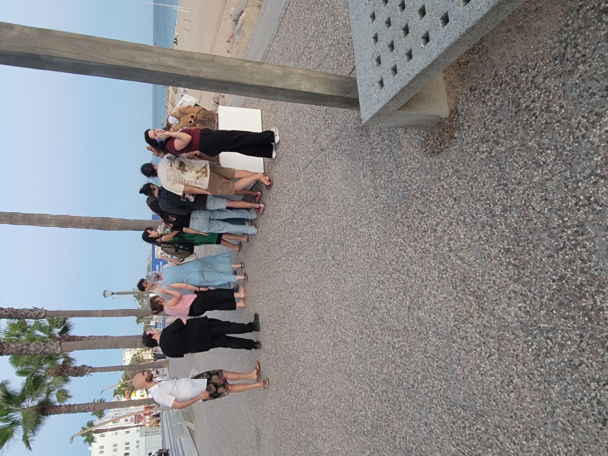 This image captures a scene along the Quarteira promenade in Portugal. Several people are gathered, some looking at an art installation situated on a white plinth. The installation appears to be a sculpture made of wood. A woman on the right is talking on her phone. In the background, palm trees line the promenade, and beyond that, the sandy beach and the Atlantic Ocean are visible. The ground is paved with a textured, speckled surface. A portion of a modern, concrete bench with a grid pattern is visible in the lower right of the frame. The overall lighting suggests it is either early morning or late afternoon.