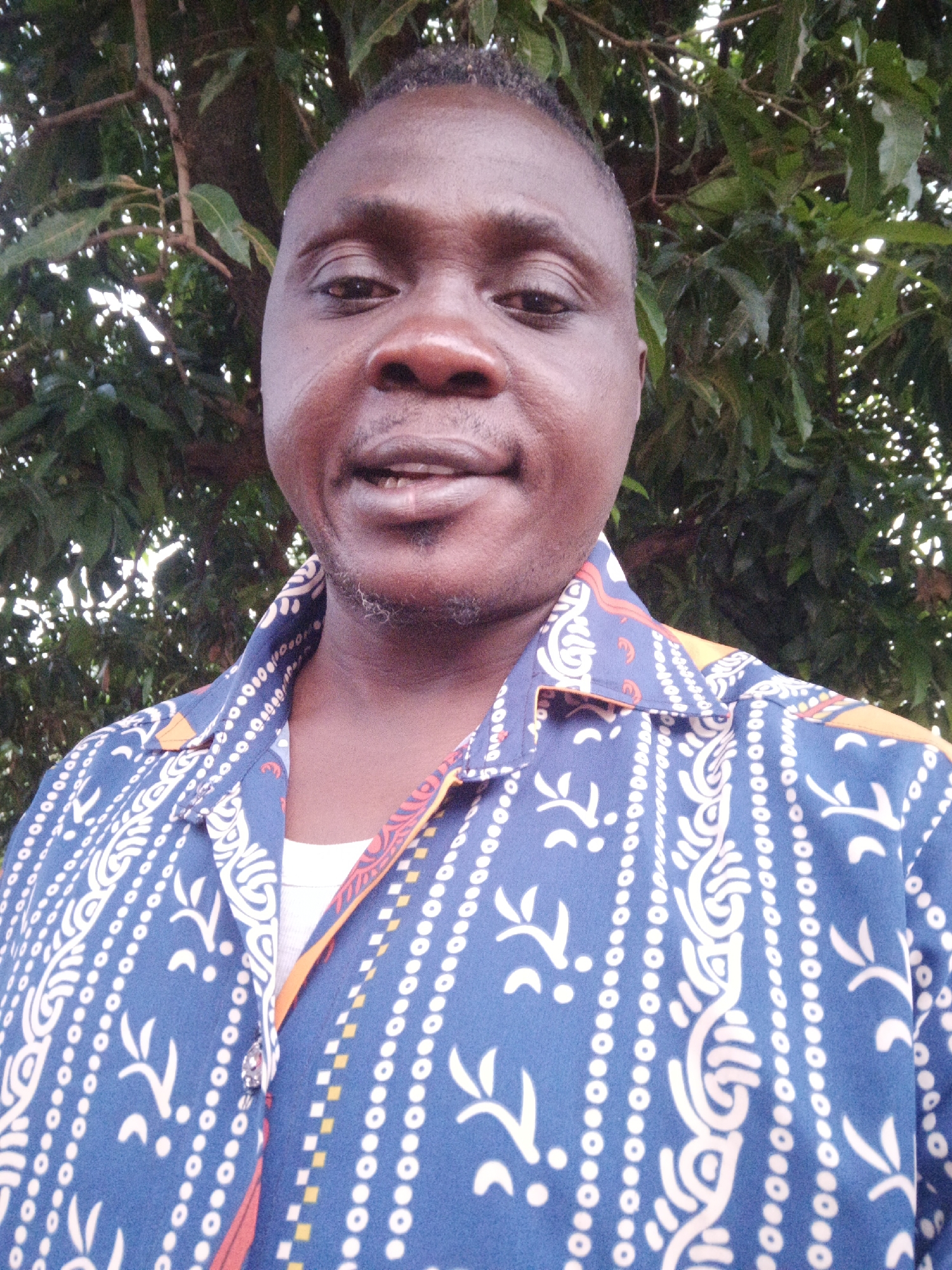 A man stands outdoors, likely in Akwanga, Nigeria, evident from visual cues possibly linked to regional fashion or vegetation. He is centered in the frame, wearing a blue patterned shirt over a white undershirt. The background features green foliage, suggesting a natural setting. The image has natural lighting.