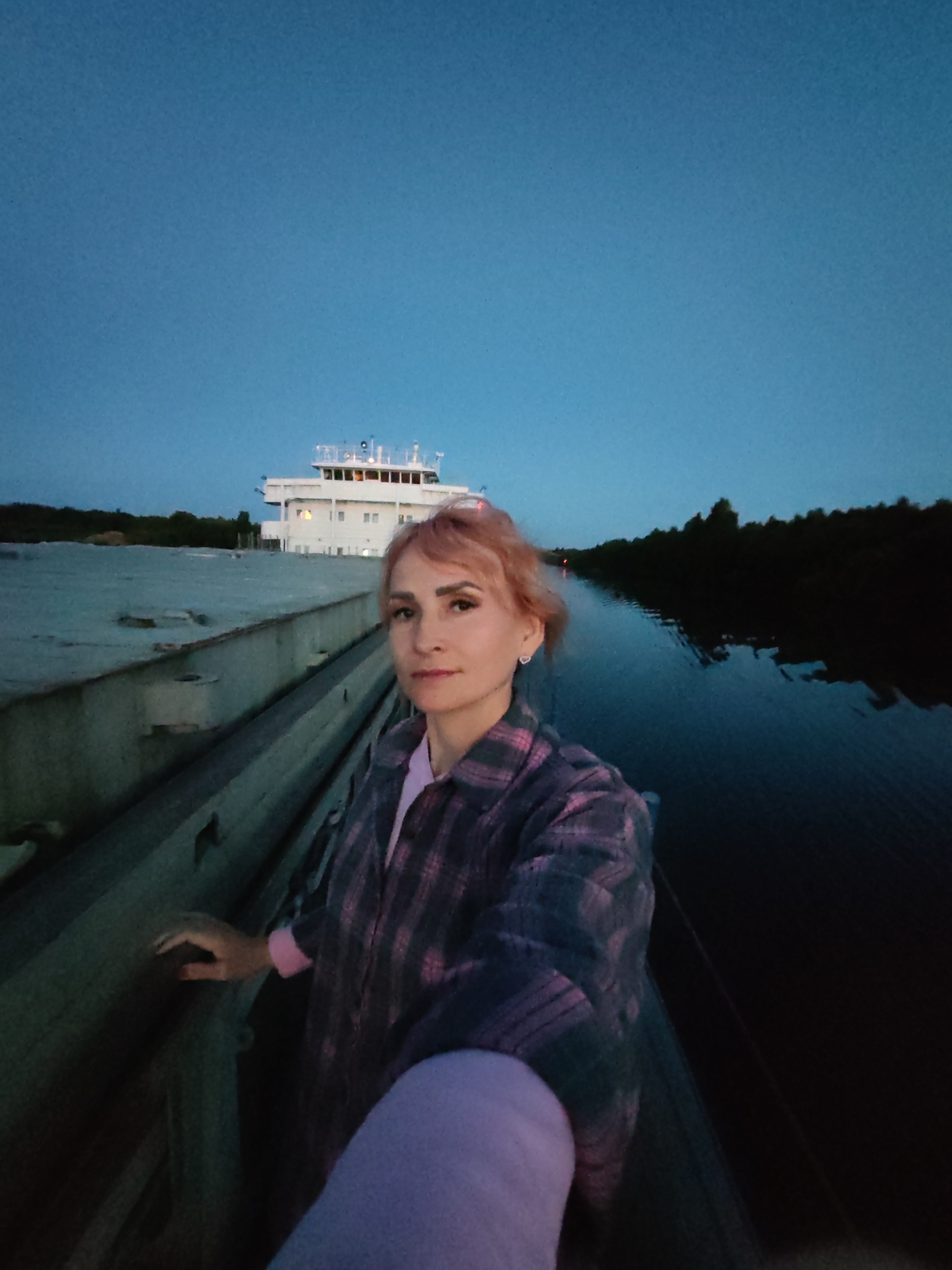 A woman with pinkish-blonde hair takes a selfie on the deck of a vessel at dusk. She is wearing a plaid shirt over a pink top. Behind her, a large white ship with multiple decks and windows is visible. The scene appears to be on a body of water, possibly a river or canal, with trees lining the banks on the right side of the image. The sky is a deep blue, indicating twilight. The location is Annensky Most, Russia.
