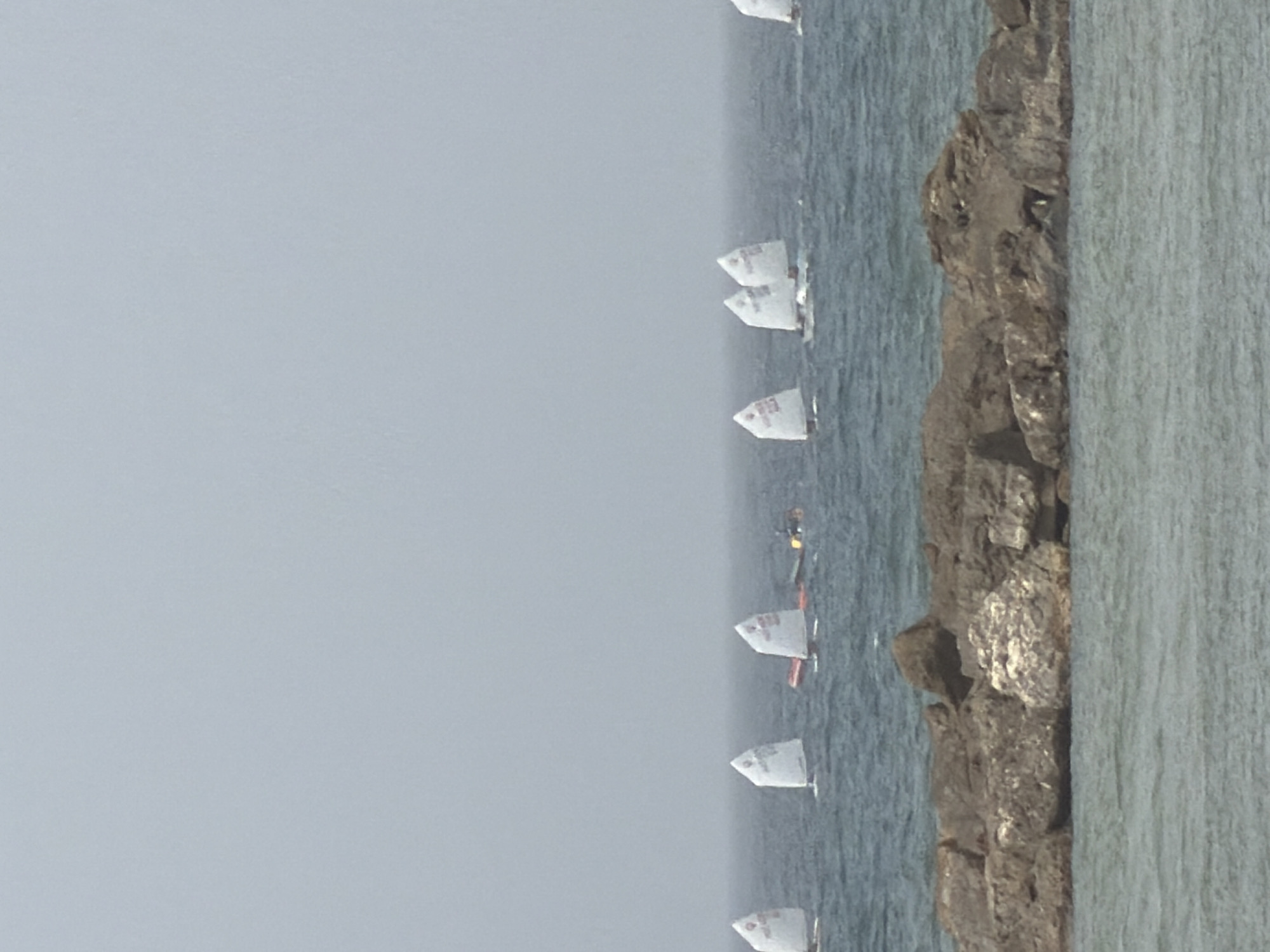 The image shows a coastal scene, possibly of Tel-Aviv, Occupied Palestine. The sea occupies the lower right portion of the frame, with a rocky shoreline separating it from the water. On the left, there is a cloudy sky. Several small sailboats with white sails are visible in the water. Between the sailboats and the rocky shoreline, a person on what appears to be a kayak is also visible.