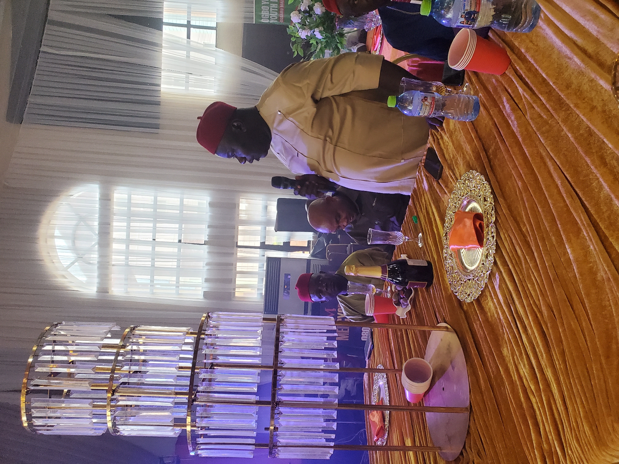 The image depicts a meeting or event taking place indoors. The main focus is a man, presumably giving a speech, who is wearing a light-colored outfit and a red cap. He is holding a microphone in his left hand. Other individuals are seated at a table, some wearing similar red caps. The table is covered with a gold-colored, draped fabric and adorned with water bottles, cups, and decorative elements. A chandelier-like structure with crystal-looking components hangs prominently in the lower-left of the frame. The background features window treatments that partially obscure the exterior. There is also a board with “OGBO YOUTH” and other words. The location is Unknown, Unknown.