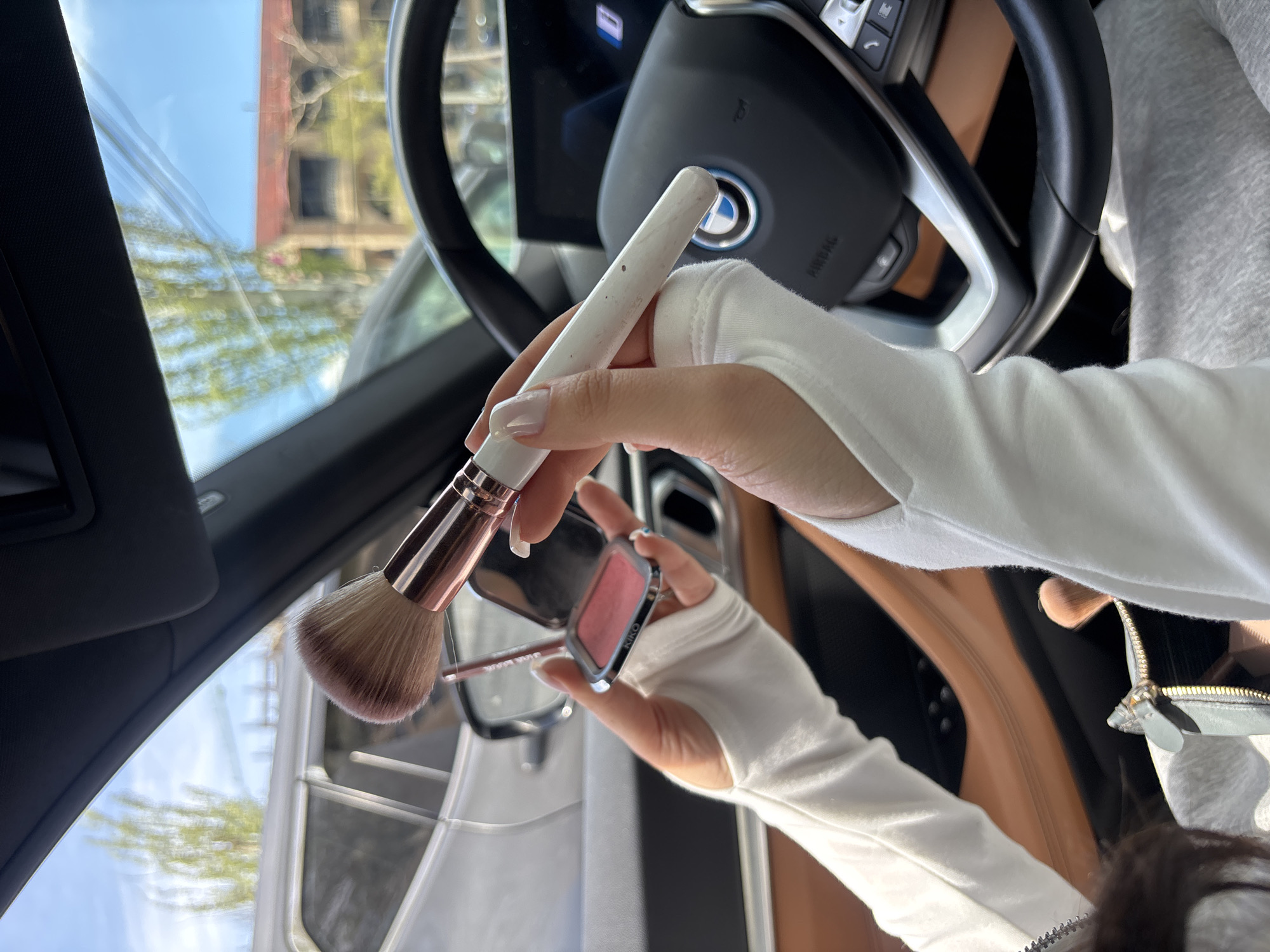 A woman is applying makeup in a BMW. She is holding a blush brush in her right hand and a blush compact in her left. The car has a black steering wheel with the BMW logo and tan interior. The woman is wearing a white, long-sleeved shirt or jacket with thumbholes. The background shows a blurry cityscape through the car window. The location is Yerevan, Armenia.