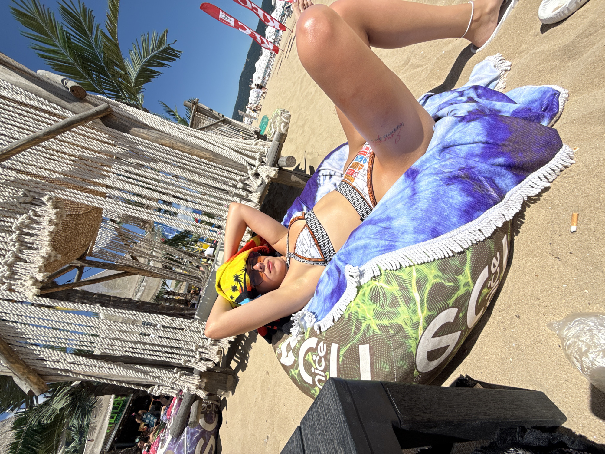 A young woman with a towel on her head and sunglasses is lying on a beanbag chair on a sandy beach. She is wearing a bikini. A colorful towel is draped over her. The background shows a beach with people, umbrellas, and palm trees. In the distance, there are buildings and hills. The scene appears to be a sunny day at a beach resort, likely in Nesebar, Bulgaria, given the context.