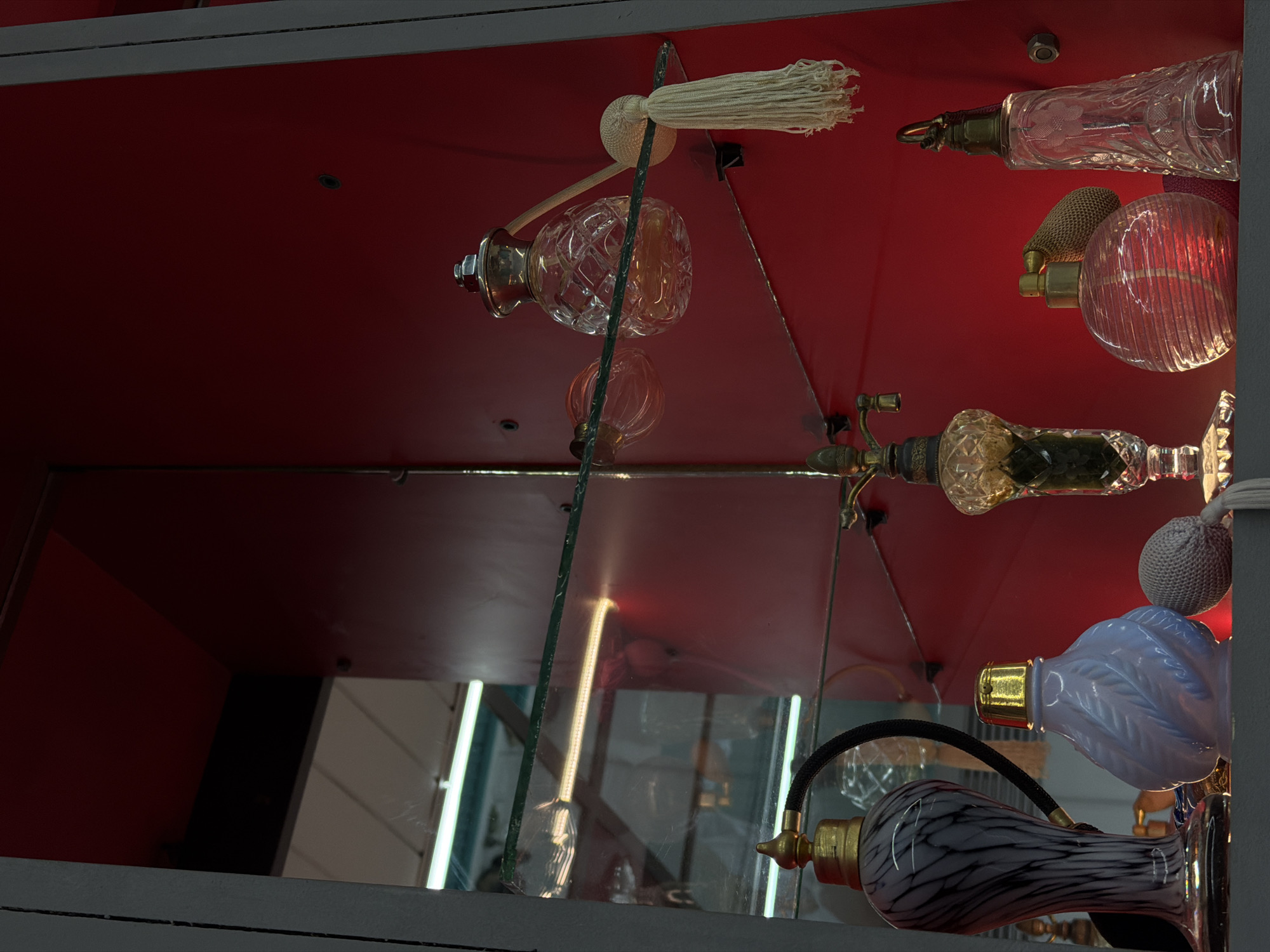 The image displays a collection of antique and vintage-style perfume bottles arranged on two shelves within a display cabinet, located in a commercial or exhibition establishment in Yerevan, Armenia. The cabinet features a deep red background, reflecting ambient light. Dark grey vertical frames border the display.
On the upper glass shelf, positioned against the red background, a clear, faceted crystal perfume bottle with a silver-toned atomizer cap contains an amber-colored liquid. Attached to it is a spherical, white netted atomizer bulb with a matching tassel hanging below. To its left, a smaller, clear amber-tinted bottle is visible. Reflections of overhead fluorescent lighting are cast onto the glass shelf.
On the lower, dark grey shelf, from left to right:
1. A tall, slender perfume bottle with a grey and white marbled glass design, topped with a brass-colored atomizer pump and a black braided hose.
2. A light lavender-colored opaque glass bottle with a raised leaf-like pattern, featuring a gold-toned cap. A grey netted atomizer bulb rests beside it.
3. A taller, ornate dark amber or green glass bottle on a pedestal base, topped with an intricate brass-colored finial.
4. A spherical, clear glass bottle with horizontal ribbing, containing a reddish liquid, fitted with a brass-colored atomizer pump and a dark netted atomizer bulb.
5. A tall, conical clear glass bottle with etched floral patterns, topped with a brass-colored stopper from which a red cord extends.
The scene is static, devoid of people, and highlights the varied designs and materials of the perfume bottles.
