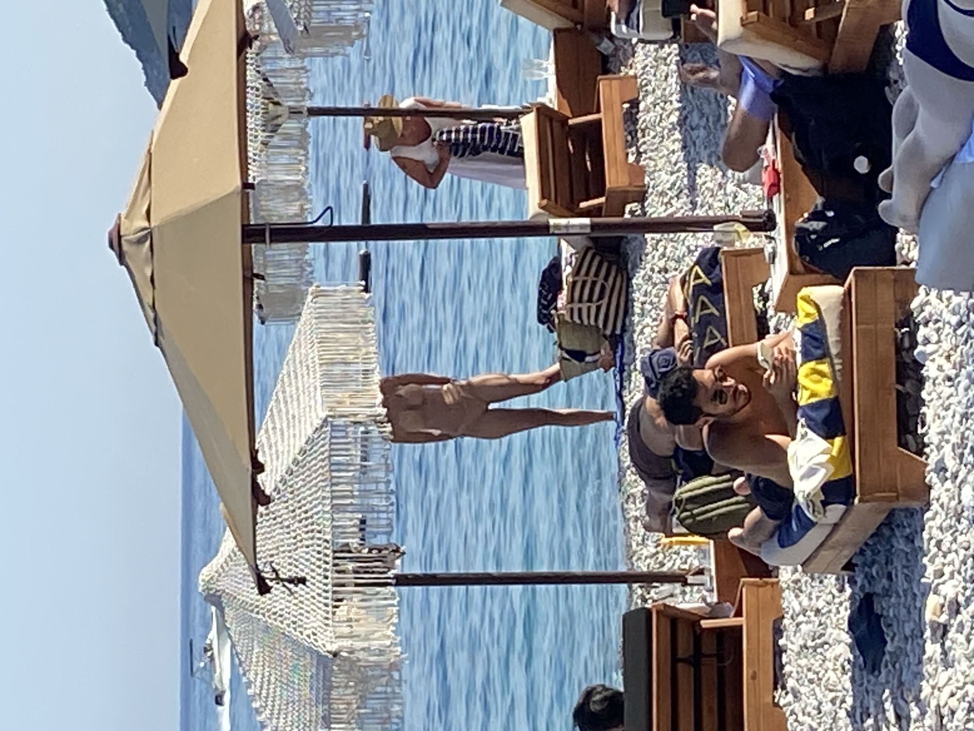 The image shows a sunny day at a pebbled beach in Prijevor, Montenegro, with the Adriatic Sea in the background. Several beach chairs are arranged on the shore. In the foreground, a man is lying on a beach chair, looking at his phone. He is wearing sunglasses and dark shorts. To his left, another person is lying on a beach chair, their face obscured. Further back, a person in a light-colored swimsuit is standing, their upper body obscured by a beach umbrella. A woman wearing a white dress and a straw hat is standing near the water, holding a striped towel. Beach umbrellas with decorative fringe are providing shade. A boat is visible in the distance on the calm blue water. The ground is covered in small, smooth pebbles.