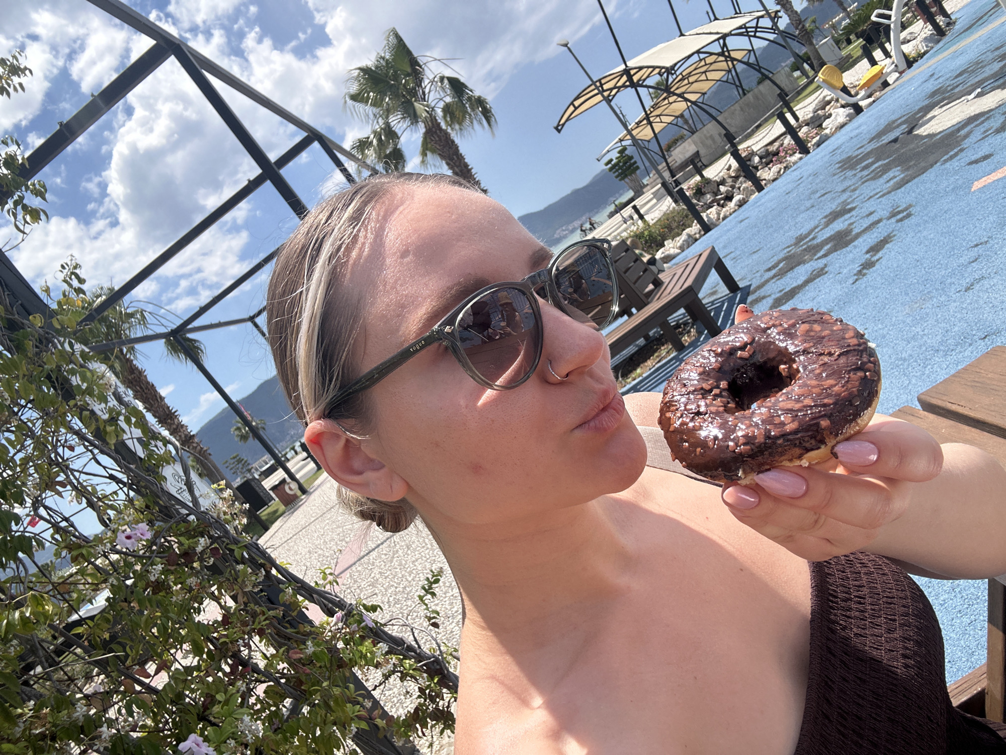 A woman is holding a chocolate donut in the city of Fethiye, Turkey. The woman has her hair pulled back in a bun and is wearing sunglasses. She is standing outside near what appears to be a small amusement park area and is holding the donut near her face while facing the camera. There is a palm tree and other greenery nearby. The sky is blue with scattered clouds. The amusement park area has a blue surface and there are benches and other structures in the background.