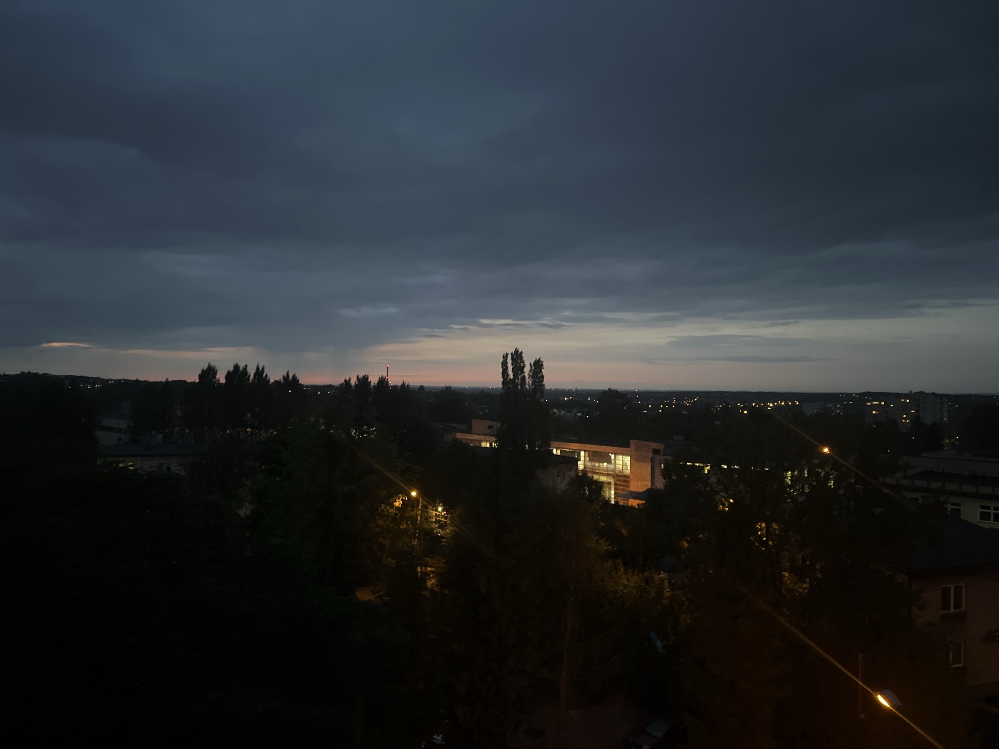 The image captures an urban landscape at dusk from an elevated viewpoint. The sky is a deep, cloudy gray, with a faint gradient of orange and pink along the horizon, suggesting the last rays of sunlight. Below the sky, a dark silhouette of trees lines the horizon, interspersed with buildings. Several trees are tall and pointed, resembling poplars.
In the mid-ground, illuminated buildings are visible, with one structure in particular featuring large windows that glow with a warm, artificial light. This building appears modern with geometric lines. Streetlights dot the scene, casting pools of yellow light on the pathways and illuminating some of the surrounding trees. The foreground is dominated by darkness, with some faint outlines of vegetation and potentially debris. The overall impression is one of a city settling into evening, with artificial lights beginning to counter the natural dimness. There are no people or discernible human activity visible in the image. The location is described as Unknown, Unknown.
