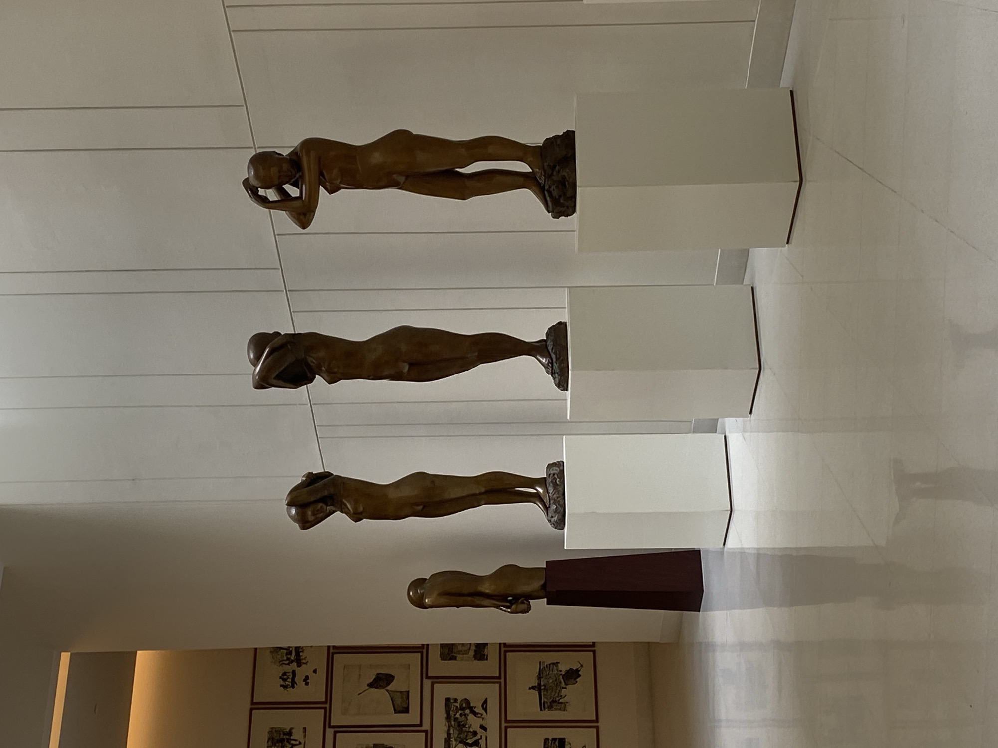 The image depicts an art installation featuring four bronze, nude, female figures in varying poses. Each sculpture is mounted on a different plinth, ascending in height from bottom to top. The plinths are either white or a darker maroon color. The background includes a white wall with horizontal indentations, and a lower section featuring framed black and white artworks on the wall.
The location is likely an art museum or gallery. While the prompt asks if the location is in Bangkok, Thailand, there is no concrete evidence to confirm this based solely on the image.