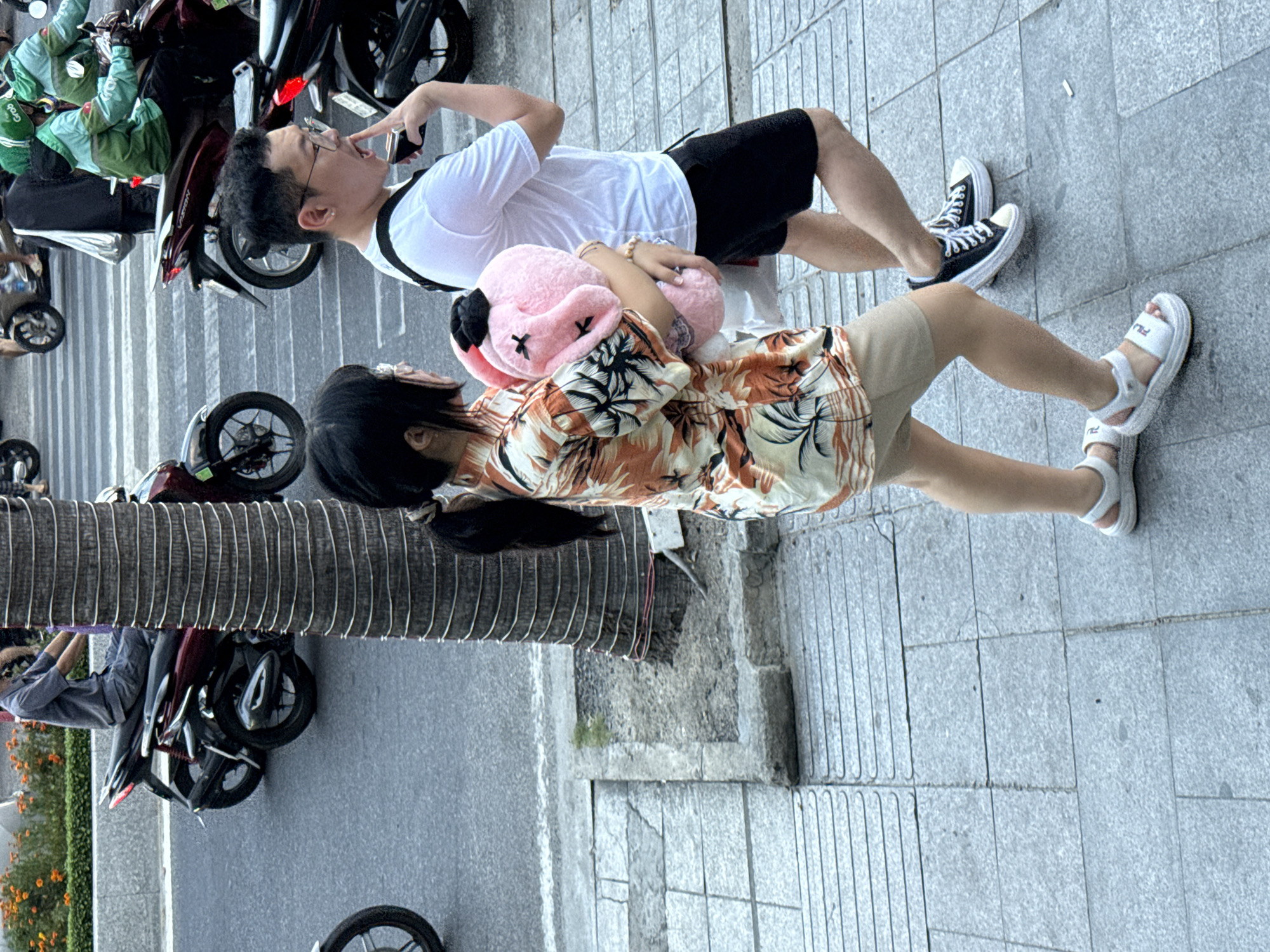 In Nha Trang Ward, Vietnam, a man and a woman are walking on a sidewalk alongside a street with traffic. The woman, on the left, wears a floral print short-sleeved shirt and tan shorts, carrying a large pink stuffed bunny. She has dark hair pulled back in a ponytail and wears white sandals. The man, on the right, wears a white t-shirt, black shorts, and black sneakers with white laces. He has glasses and is touching his finger to his mouth. In the background, motorcycles and scooters are visible on the street and crossing a pedestrian walkway. A tall, ribbed palm tree trunk is on the right side of the sidewalk.