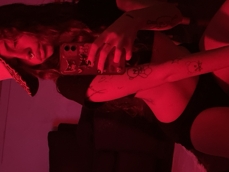 The scene depicts a single individual, likely female, in an indoor setting bathed entirely in red light. The person is positioned in a seated or kneeling posture, facing forward, with a dark, indistinct piece of furniture visible in the background to the left. She wears a dark, short-sleeved crop top and dark shorts. A dark, patterned wide-brimmed hat partially covers her head and forehead. A silver septum piercing is visible in her nose. She holds a dark-cased smartphone, adorned with cartoonish characters and heart details, with her right hand positioned in front of her face, indicating a mirror selfie. Her lips are slightly pursed. Multiple tattoos are visible on her arms, including a teddy bear outline on her right forearm and a skull-like bear outline on her left forearm. The specific location within the city of Buenos Aires, Argentina, cannot be ascertained from the visual evidence.
