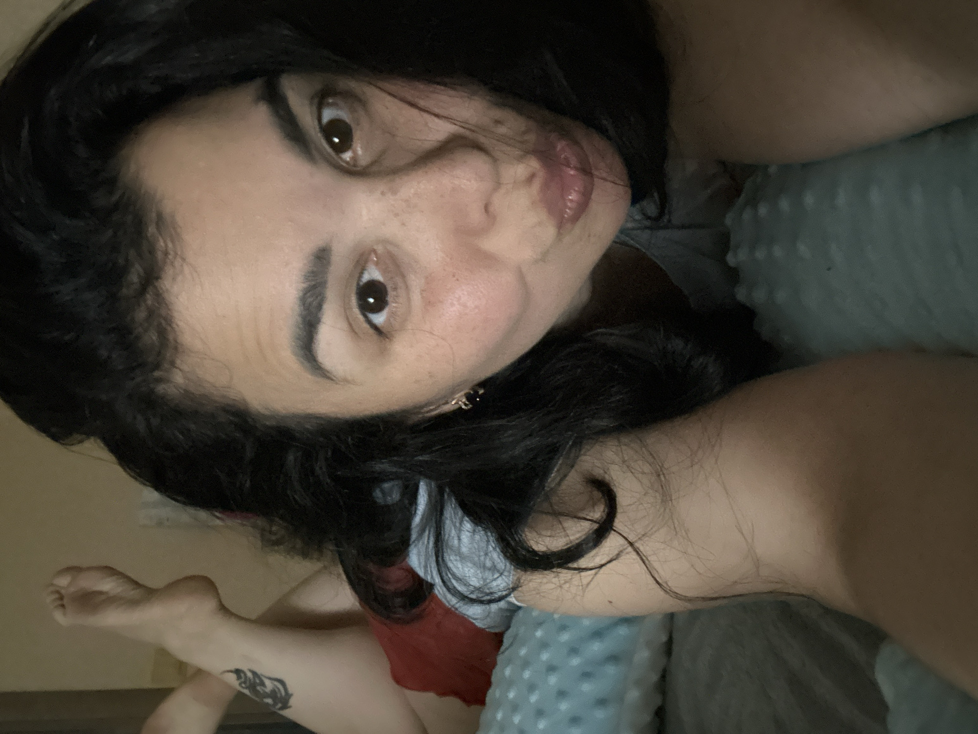 A woman with dark hair and brown eyes is lying on a bed, looking at the camera. She has freckles on her face. She is wearing a grey t-shirt and red shorts. She is lying on a grey blanket. Her leg is visible in the lower left corner of the image, with a tattoo on the lower leg. The location is unknown.