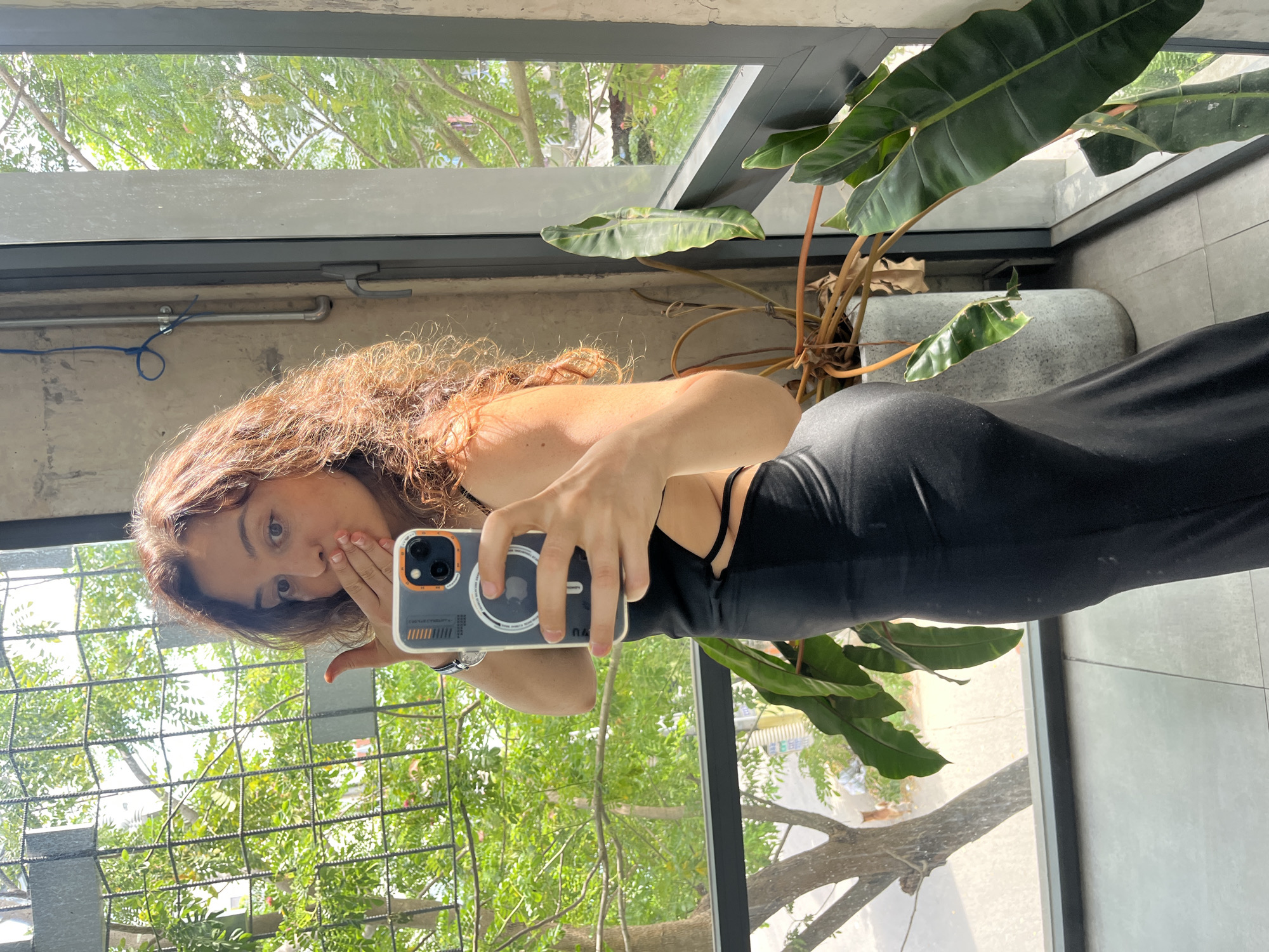 A woman stands in front of a large window, holding a smartphone to take a mirror selfie. She has long, curly brown hair and is wearing a black dress with cutouts on the sides. She is blowing a kiss towards the camera. Outside the window, lush green trees and a city street are visible. The interior appears to be a modern space with concrete walls and large windows. The presence of trees and the overall architecture suggest an urban environment, possibly within Phu Thuy Ward, Vietnam, given the context.