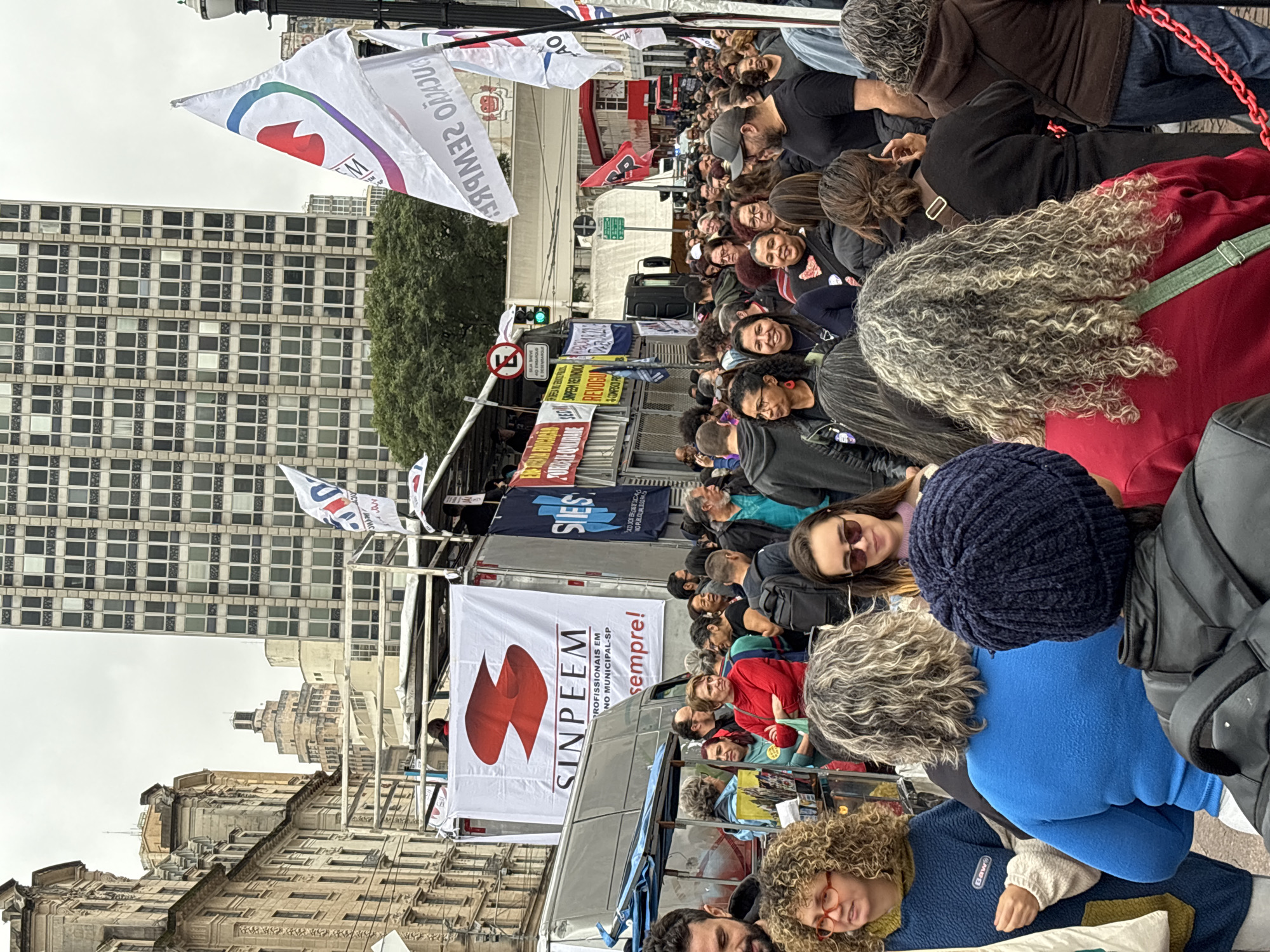 A daytime, eye-level shot portrays a sizable crowd in what appears to be a protest or demonstration in the city of São Paulo, Brazil. The crowd is tightly packed, with individuals of various ages and appearances. Many are looking in different directions, while some seem to be interacting with each other.
A truck serves as a makeshift stage, draped with banners that read "SINPEEM PROFISSIONAIS EM NO MUNICIPAL-SP sempre!" and "SIESP" along with other posters and flags bearing political messages. The signs and banners indicate the demonstration pertains to professional issues.
The urban setting is defined by buildings, a street, and a traffic light. Old, colonial-style buildings are visible, juxtaposed against a modern, high-rise structure in the background. The architecture suggests this could be the historic center of São Paulo.