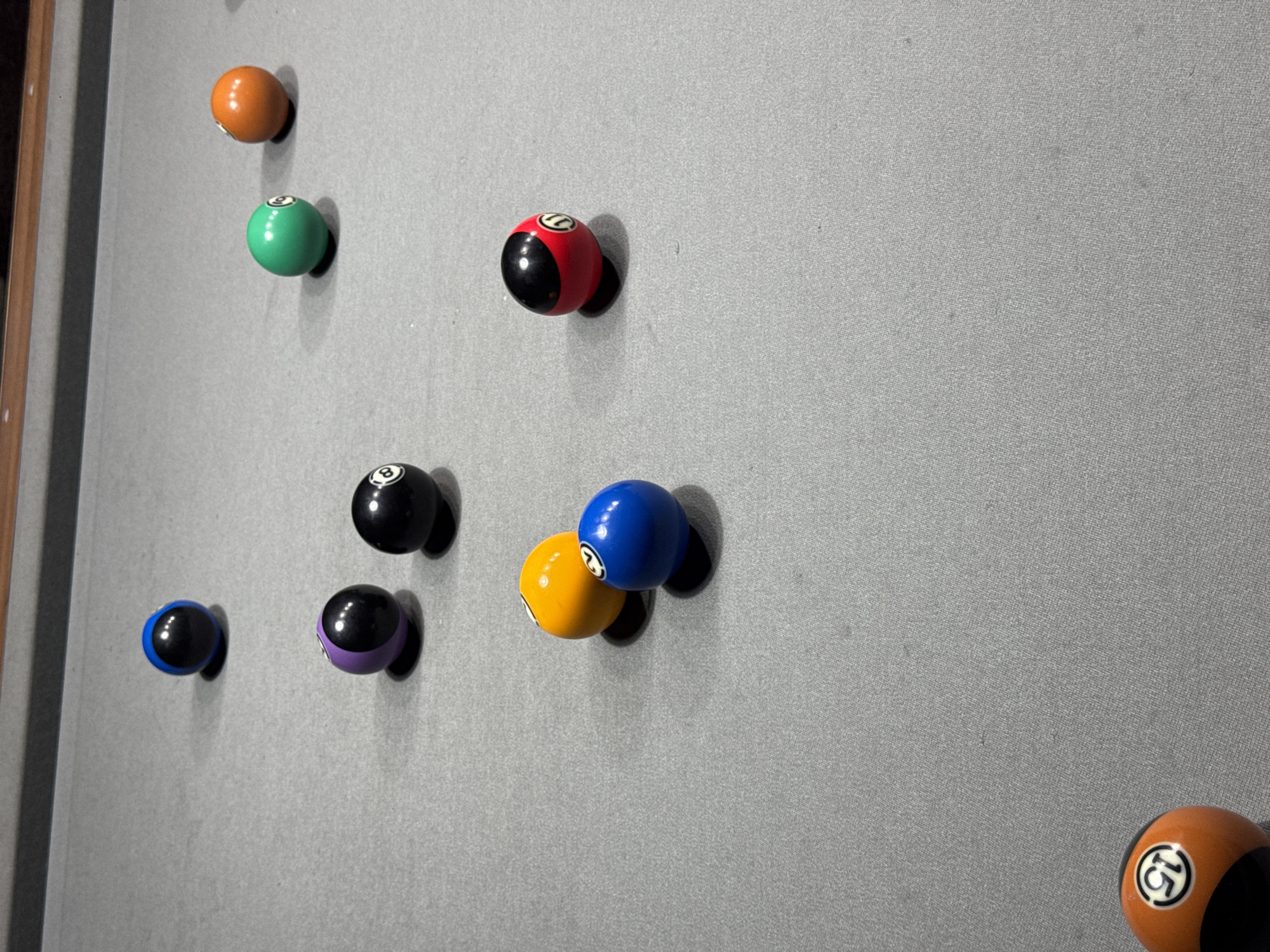 An overhead, eye-level, medium shot captures several billiard balls on a grey felt pool table. The balls are arranged in a scattered pattern across the table, with some closer to the foreground and others further back. In the foreground, a partially visible orange and black billiard ball, numbered 15, rests at the bottom left of the frame. Moving towards the center, a cluster of three balls includes a yellow ball, a blue ball numbered 2, and a red ball numbered 11 with a black top. To the left of this cluster, a purple ball numbered 3 sits next to a black 8-ball. Further back and to the left, a blue ball with a black top is positioned. In the upper right quadrant of the image, a green ball numbered 6 is situated near an orange ball numbered 7. A wooden rail runs along the top edge of the image, suggesting the boundary of the pool table. The lighting appears to be artificial, casting soft shadows from the balls onto the felt. The scene is indoors, and there are no people visible. The image is a still photograph with no discernible action or interaction. No specific location within Burgas, Bulgaria, can be determined from the visual information provided.