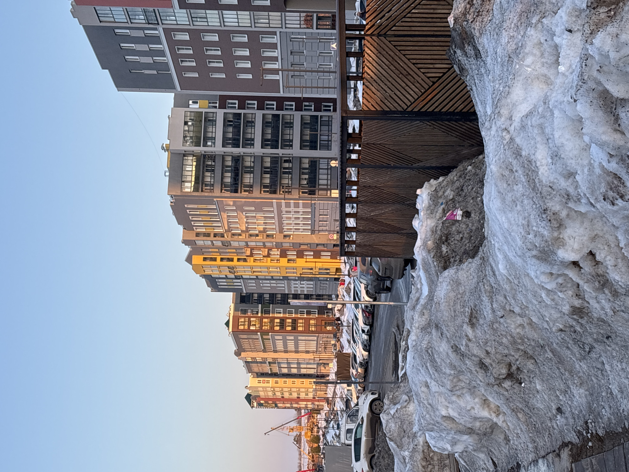 This image depicts an urban street scene in Pervomaysky, Russia, under clear, bright skies, likely during late afternoon or early morning based on the low-angle sunlight. The foreground features large, irregular mounds of dirty, partially melted snow and ice alongside a paved sidewalk on the left. Scattered debris is visible within the snow.
A row of multi-story residential buildings, varying in height and featuring diverse architectural styles and color palettes (including shades of grey, brown, red, and yellow), lines the street on the right, extending into the background. Many buildings have visible windows and balconies. A wooden fence with a diagonal slatted pattern stands prominently in the mid-ground on the right, partially obscuring the street.
The asphalt street is lined with numerous parked vehicles, predominantly sedans and SUVs, with a white van also visible on the left. The vehicles are arrayed in parking spots along the curb. In the distant background on the far left, construction cranes are visible against the horizon, indicating ongoing development. A solitary, dark-clothed individual is faintly visible behind the wooden fence in the mid-ground on the far right, partially obscured by a light pole. No specific actions or interactions are clearly discernible.