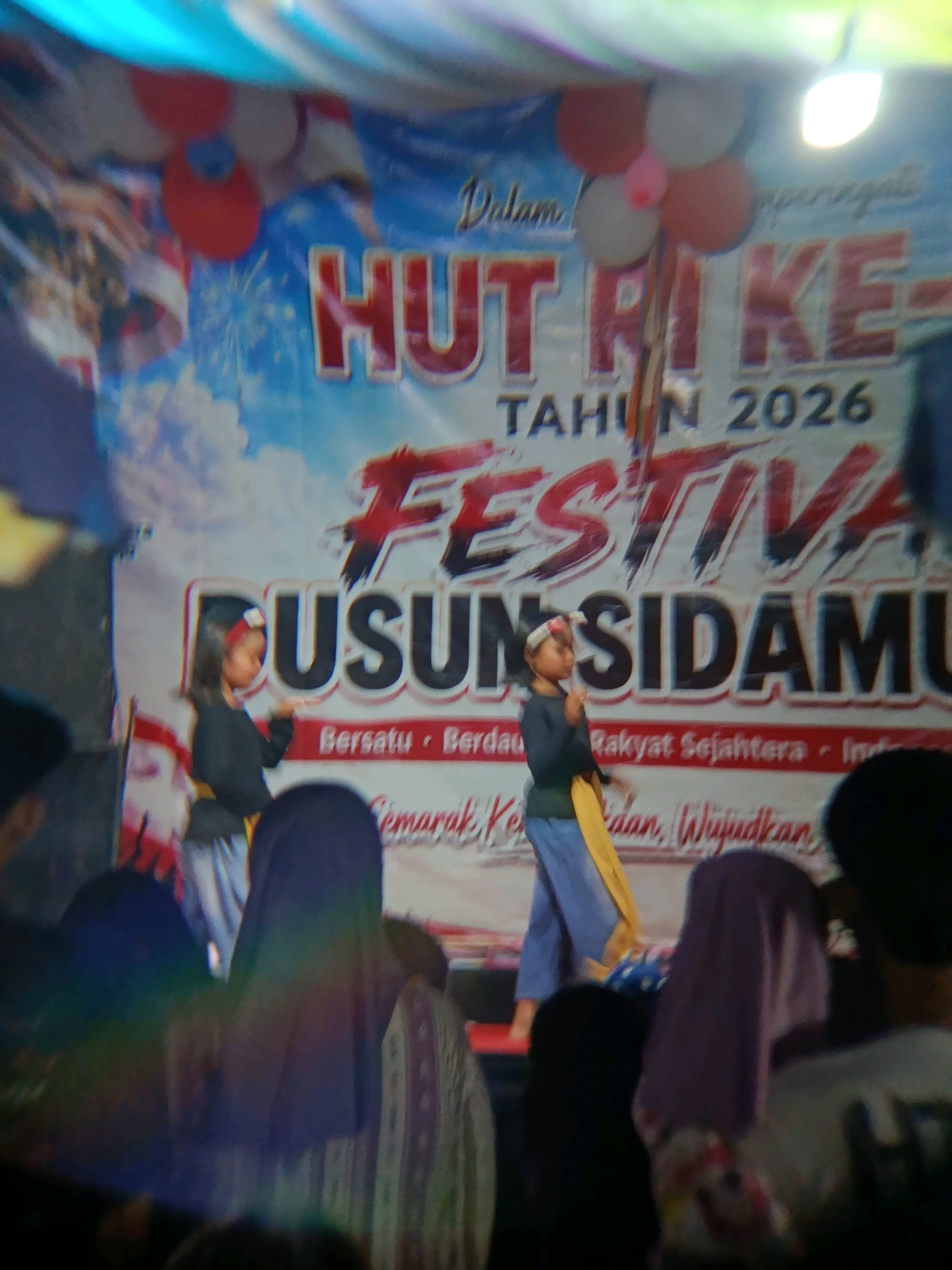 In a dimly lit indoor venue, possibly a community hall or stage in Binangun, Indonesia, a banner reads "HUT RI KE- TAHUN 2026 FESTIVA GUSUN SIDAMU" with text below including "Bersatu - Berdaulat - Rakyat Sejahtera - Ind." and "Semarak, Kebangsaan, Wujudkan". Two children, one in a black top and blue pants with a red headband, and another in a dark long-sleeved shirt and blue pants with a yellow sash, are performing on a stage. They are positioned in front of the banner, with the child in the yellow sash on the right. In the foreground and background, several audience members are visible, some with heads covered in hijabs. The lighting appears to be from above and to the right, creating some lens flare and blurring. The overall atmosphere suggests a local celebration or cultural event.