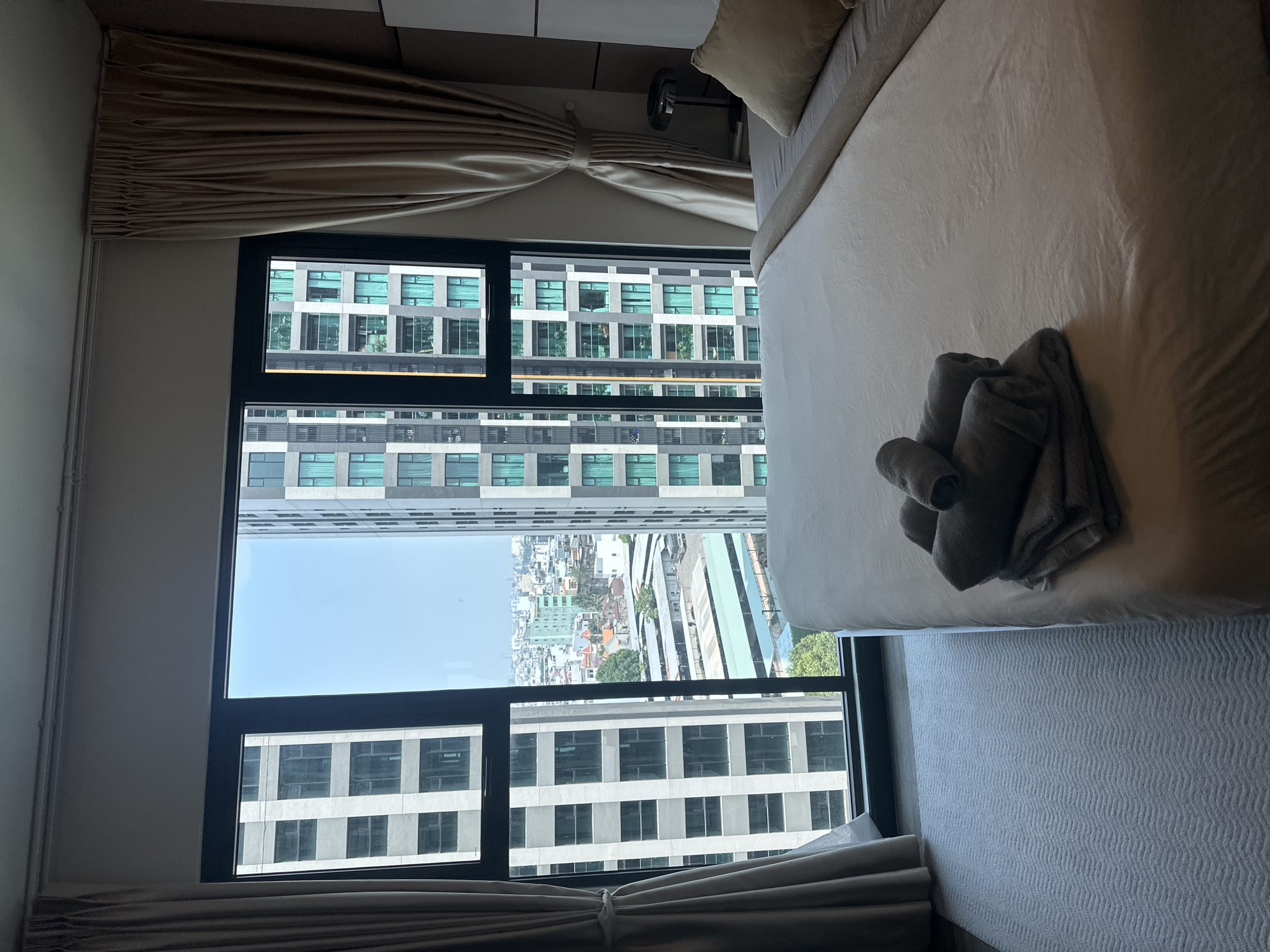 The image depicts a bedroom interior looking outward through a large, multi-pane window in Thủ Đức, Vietnam. The foreground shows a bed with a white duvet, a beige throw blanket, and a beige pillow. Stacked on the bed, near the foot, are three grey towels, two rolled and one folded flat. To the left of the bed, a light grey rug with a subtle chevron pattern covers the floor up to the window frame. Beige curtains hang on either side of the window, partially drawn and tied back on the left, obscuring a small portion of the view. A dark-colored, modern table lamp is visible on a nightstand to the right of the bed.
Through the window, a dense urban landscape is visible under a clear blue sky. Two prominent high-rise buildings frame the central view: one with a light grey facade and numerous windows on the left, and another with teal-tinted windows and balconies on the right. In the middle distance, between these towers, a lower-rise urban sprawl includes various buildings, some with red or terracotta roofs, indicative of residential or commercial structures. Below, an expansive area features several long, low-rise structures with corrugated roofs, appearing to be a parking facility or an industrial compound. No individuals are present in the scene.