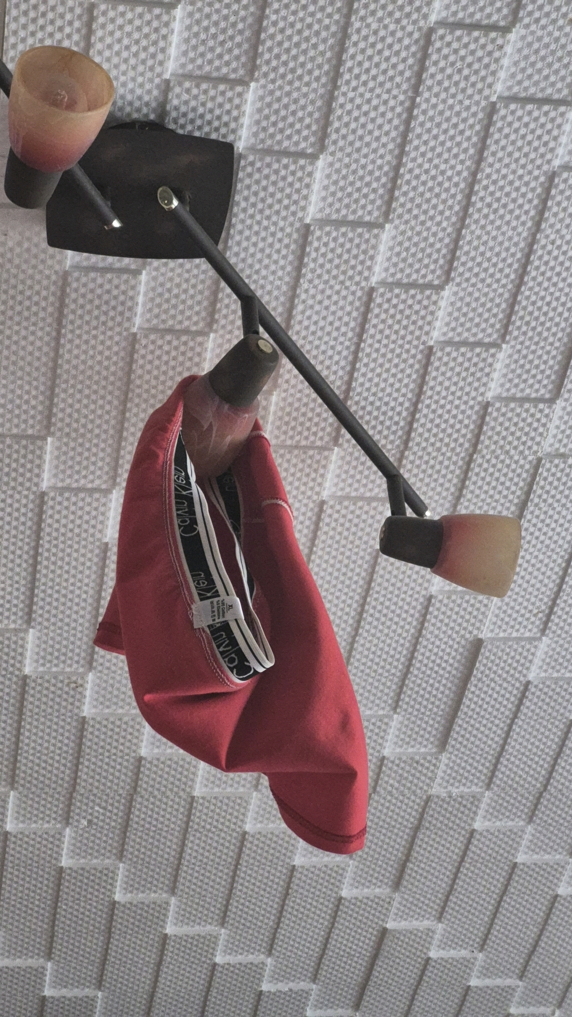 A red garment, possibly underwear based on the visible waistband with "Calvin Klein" branding, hangs from a two-armed, dark metal fixture attached to a textured white ceiling. The fixture has two glass lampshades, one of which is partially visible and emits a soft, pinkish-orange light. The ceiling's surface is composed of repeating, raised square patterns. The overall impression is of a ceiling light fixture with an item of clothing casually draped over it, likely indoors. The location is not specified in the image.