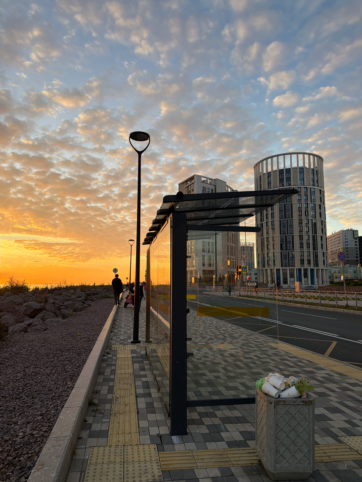 The image captures a scene at a bus stop on a waterfront promenade in Saint Petersburg, Russia, during sunset. The sky is filled with dramatic clouds illuminated by the setting sun, casting an orange and yellow glow. To the left, a rocky embankment slopes down to the water. A paved walkway, featuring tactile paving for visually impaired individuals, runs alongside the promenade.
The bus stop itself is a modern structure with a glass enclosure and a metal roof. Adjacent to the bus stop, a concrete trash bin overflows with discarded paper cups and plastic. In the background, contemporary apartment buildings with unique architectural designs, including a distinctive cylindrical tower, dominate the skyline. The street beside the promenade has visible lane markings and a yellow line, indicating it is a road.
Several people are present on the promenade. One individual walks away from the camera, accompanied by a child on a small ride-on toy. Other figures are visible further down the walkway, also appearing to be pedestrians. A lamppost stands tall next to the bus stop, and another is visible further down the promenade. Traffic lights and road signs, including a stop sign, are present on the street. The overall impression is of a well-developed urban area by the sea, bathed in the warm light of dusk.