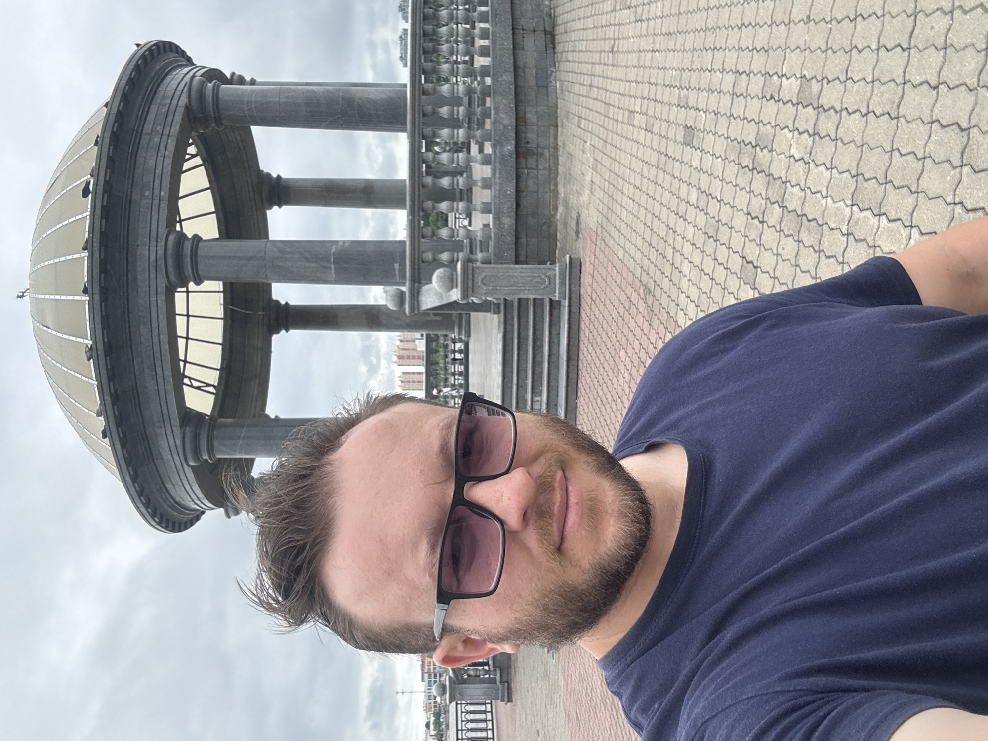 A man wearing a dark blue t-shirt and dark-rimmed sunglasses is taking a selfie in front of a large, gray, circular gazebo with a domed roof. The gazebo is supported by thick, stone pillars and has a decorative stone railing around it. Steps lead up to the gazebo. The ground is paved with interlocking gray and reddish-brown bricks. In the background, across a body of water, are several tall buildings. The sky is overcast and gray. This scene is located on the embankment of the Amur River in Blagoveshchensk, Russia, near the border with China.