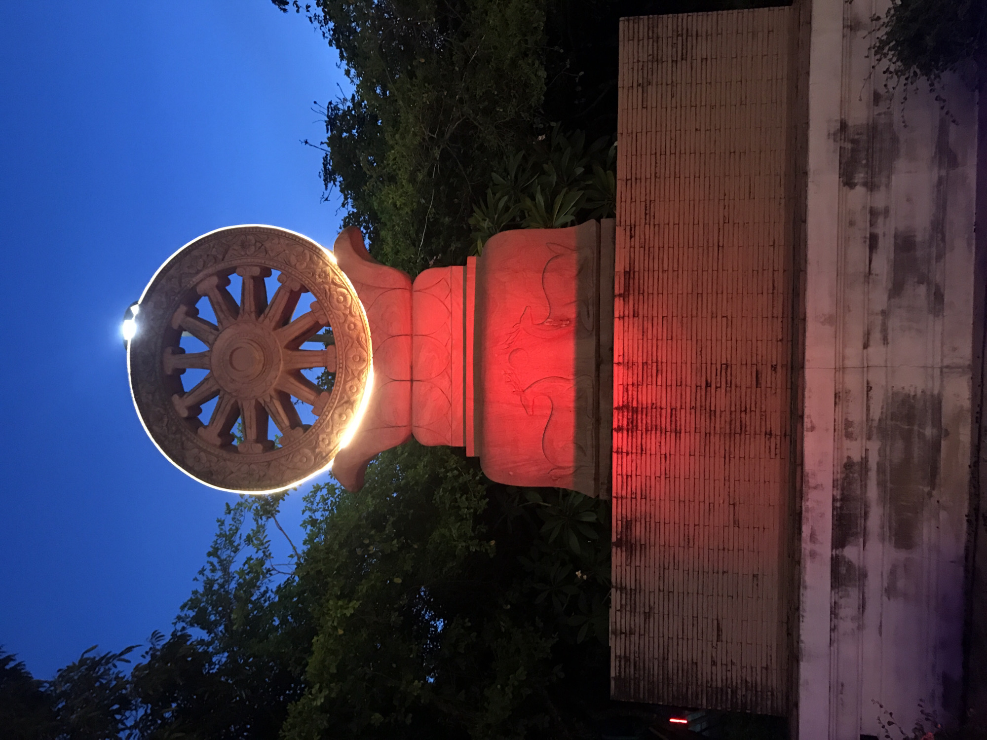 The image shows a Buddhist monument in Baan Mae Nam, Thailand, at twilight. The monument consists of a Dharma wheel, a tiered pillar with animal carvings, and a brick wall. The wheel is made of stone and illuminated by a bright light. The pillar is lit with red light, highlighting the carvings. Trees are visible in the background, and the sky is a deep blue.