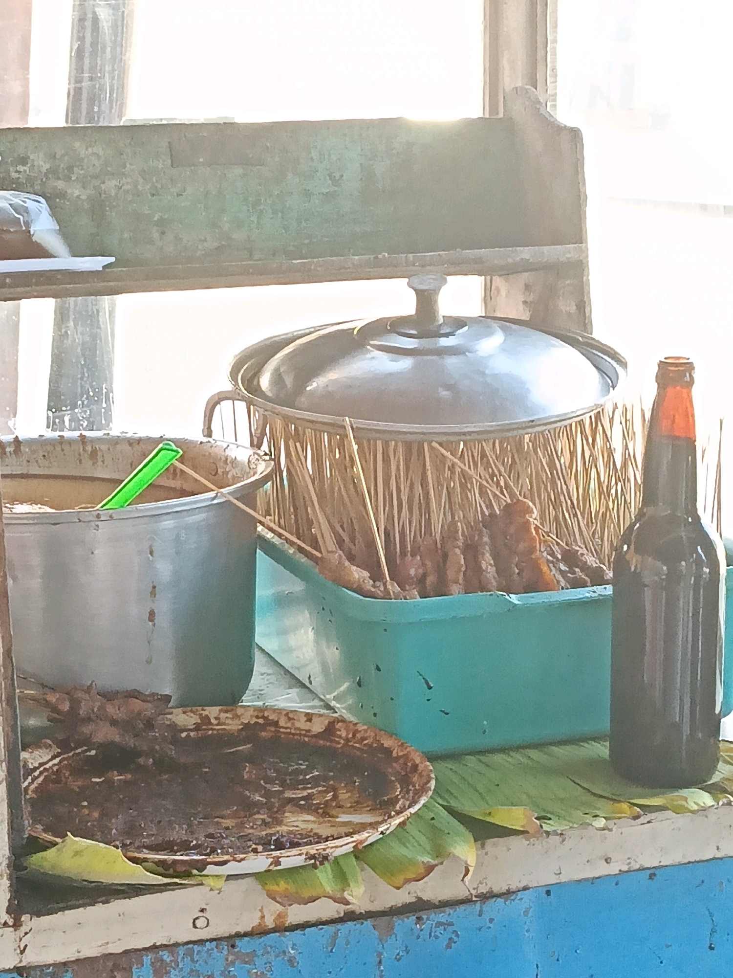 A food stall in Kedungtuban, Indonesia, displays skewers of meat arranged in a teal-colored bin covered by a metal pot lid. Next to it, a large metal pot holds a light brown liquid, with a green ladle resting inside. In front of the bin, a dirty plate with remnants of food sits on banana leaves, and a dark bottle of liquid is placed on a banana leaf. The stall is made of weathered materials with peeling paint, and the background suggests an outdoor setting with diffused sunlight.