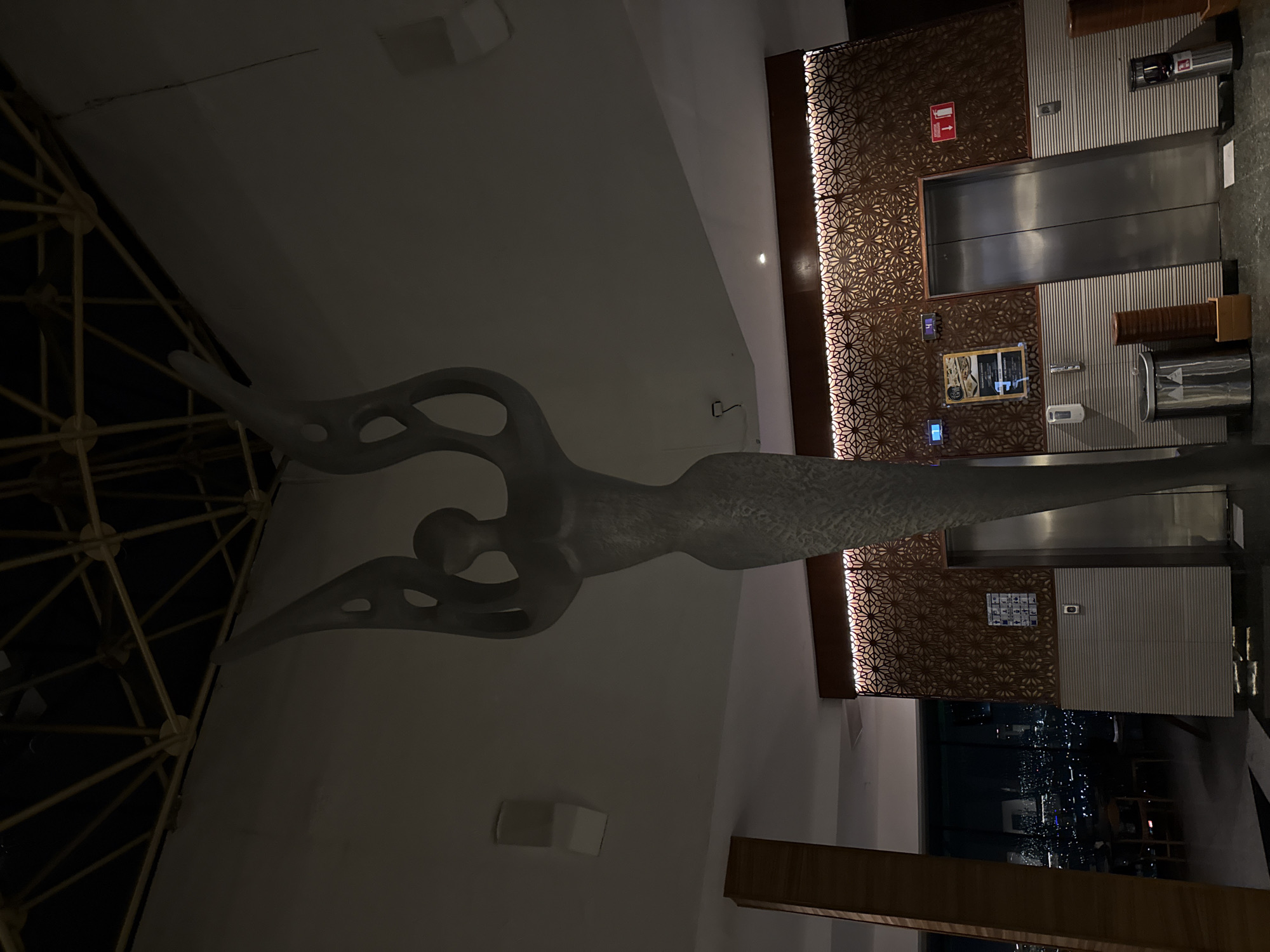 The image shows an indoor scene, likely within a building in Querétaro, Mexico, featuring modern design elements. A stylized, abstract sculpture of a human-like figure with elongated arms and hollow sections is prominently displayed. To the right, there is a textured wall panel with geometric patterns, and next to it is an elevator with stainless steel doors. Beneath the panel, a window provides a view of the cityscape at night. The lighting in the scene is subdued, highlighting the sculpture and architectural details.