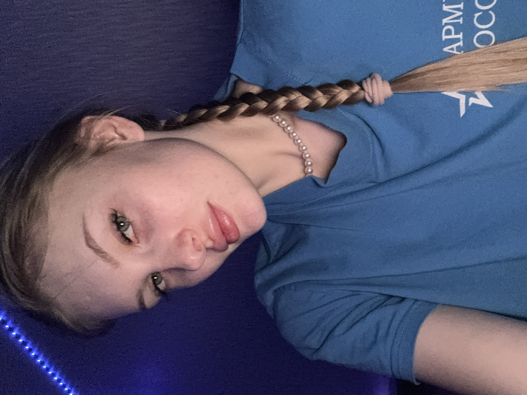 The image, rotated 90 degrees clockwise, presents a close-up of a young person lying down in an indoor setting, looking directly at the camera. The subject has light blonde or light brown hair styled in a braid that extends over their shoulder and chest, secured with a light pink or nude hair tie. They have a light complexion, green eyes, and full, slightly parted lips.
The person is wearing a short-sleeved blue t-shirt and a pearl necklace. On the upper right portion of the t-shirt, white text is partially visible, showing "APM" above a white star, and the letters "OCCO". The background is a dark blue surface, possibly a bed or mat, with a string of blue LED lights illuminating the lower left side of the frame, suggesting a nighttime or dimly lit environment. The person's expression is neutral, and their head is tilted. The scene takes place in an indoor location in Кондратово, Russia.
