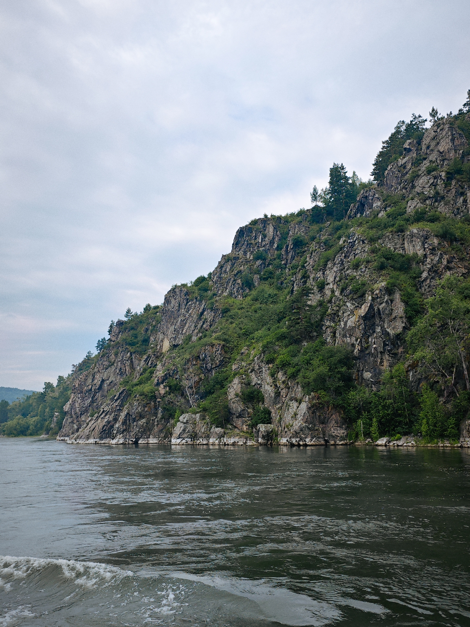 The image depicts a wide, dark river flowing in the foreground. On the right side of the image, a steep, rocky mountain rises dramatically from the riverbank, covered in lush green vegetation and scattered pine trees. The mountain's surface is rugged and uneven, with visible rock formations and crevices. The sky above is overcast with soft, grey clouds, creating a muted lighting effect. The left side of the image shows a more distant, tree-covered shoreline, suggesting a natural, undeveloped landscape. The overall scene conveys a sense of natural grandeur and remoteness. The location is the Yenisei River near the village of Atamanovo, Krasnoyarsk Krai, Russia.