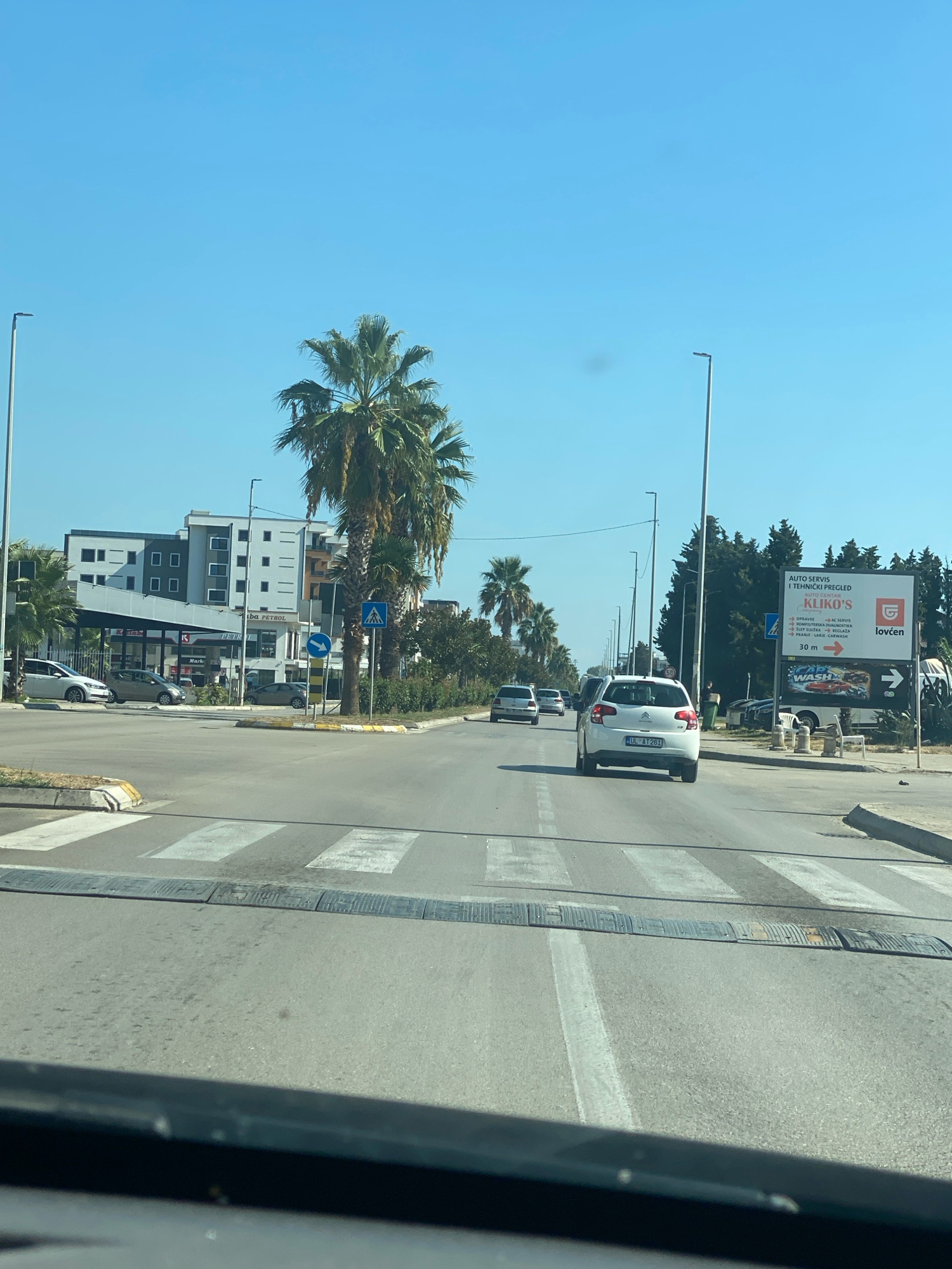 The image shows a street scene in Ulcinj, Montenegro, on a clear, sunny day. In the foreground, a car drives over a speed bump on a paved road. A pedestrian crossing is visible further ahead. In the middle ground, several cars are driving or parked on the road, and a white van is prominently featured. On the right side of the road, a large billboard advertises "AUTO SERVIS I TEHNICKI PREGLED KLIKO'S" and also features "LOVCEN OSIGURANJE" and "CAR WASH". Palm trees line the street, and a modern building complex with balconies is visible on the left. The sky is clear blue, indicating fair weather.