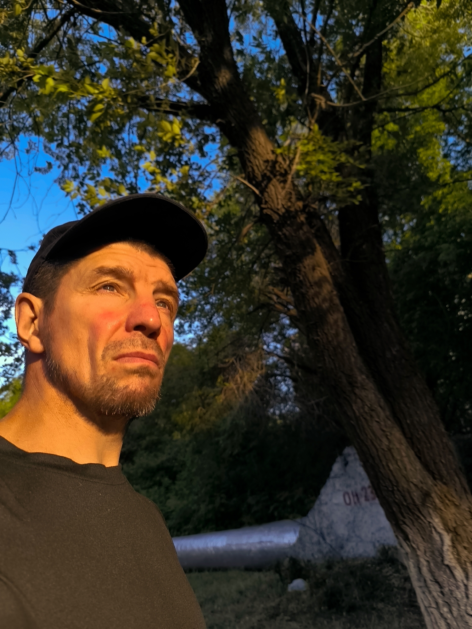 An outdoor, eye-level, medium shot captures a man wearing a black baseball cap and a black t-shirt, looking up and to the right. His face, illuminated by warm, golden hour light, shows a goatee and a slight blush on his cheeks. Behind him, a large tree trunk with textured bark dominates the right side of the frame. The background consists of dense green foliage and a bright blue sky. In the lower right quadrant of the image, partially obscured by the tree, a triangular, grey stone marker is visible with red lettering that appears to read "OH 23". To the left of the stone marker, a section of a silver, cylindrical object, possibly a pipe or conduit, runs horizontally. The scene is likely set in an area with vegetation and possibly some form of urban or industrial infrastructure, given the presence of the marker and the cylindrical object. The specific location in the city of Qarağandı city administration, Kazakhstan, is not visually evident from the image itself.