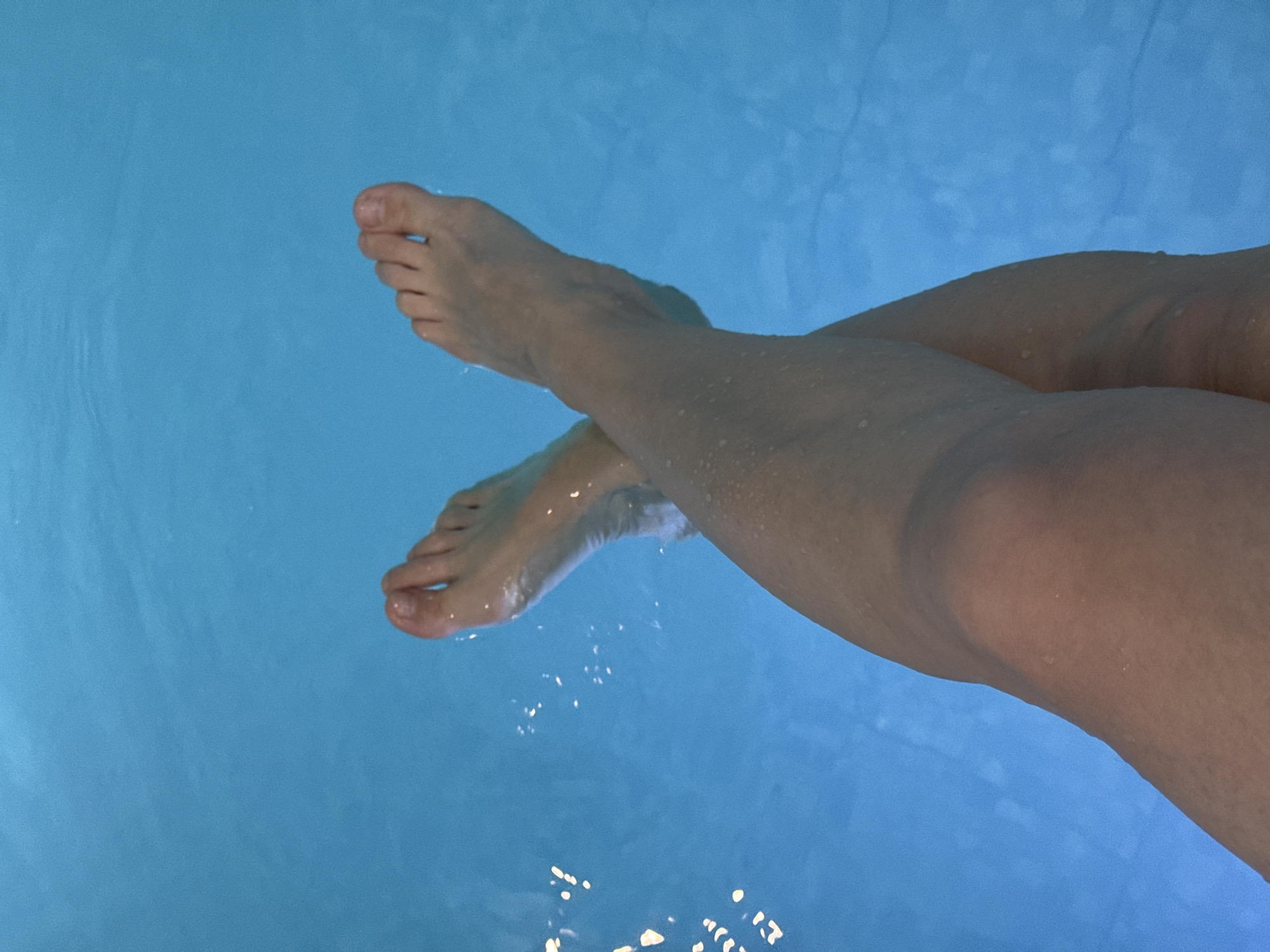 A high-angle shot depicts a person's legs and feet submerged in light blue water. The legs are crossed at the ankles. Water droplets cling to the skin of the legs. The feet are slightly submerged, causing ripples in the water. The environment surrounding the water source is unseen. There are no references to the city of Chaikovsky, Russia, in the image.