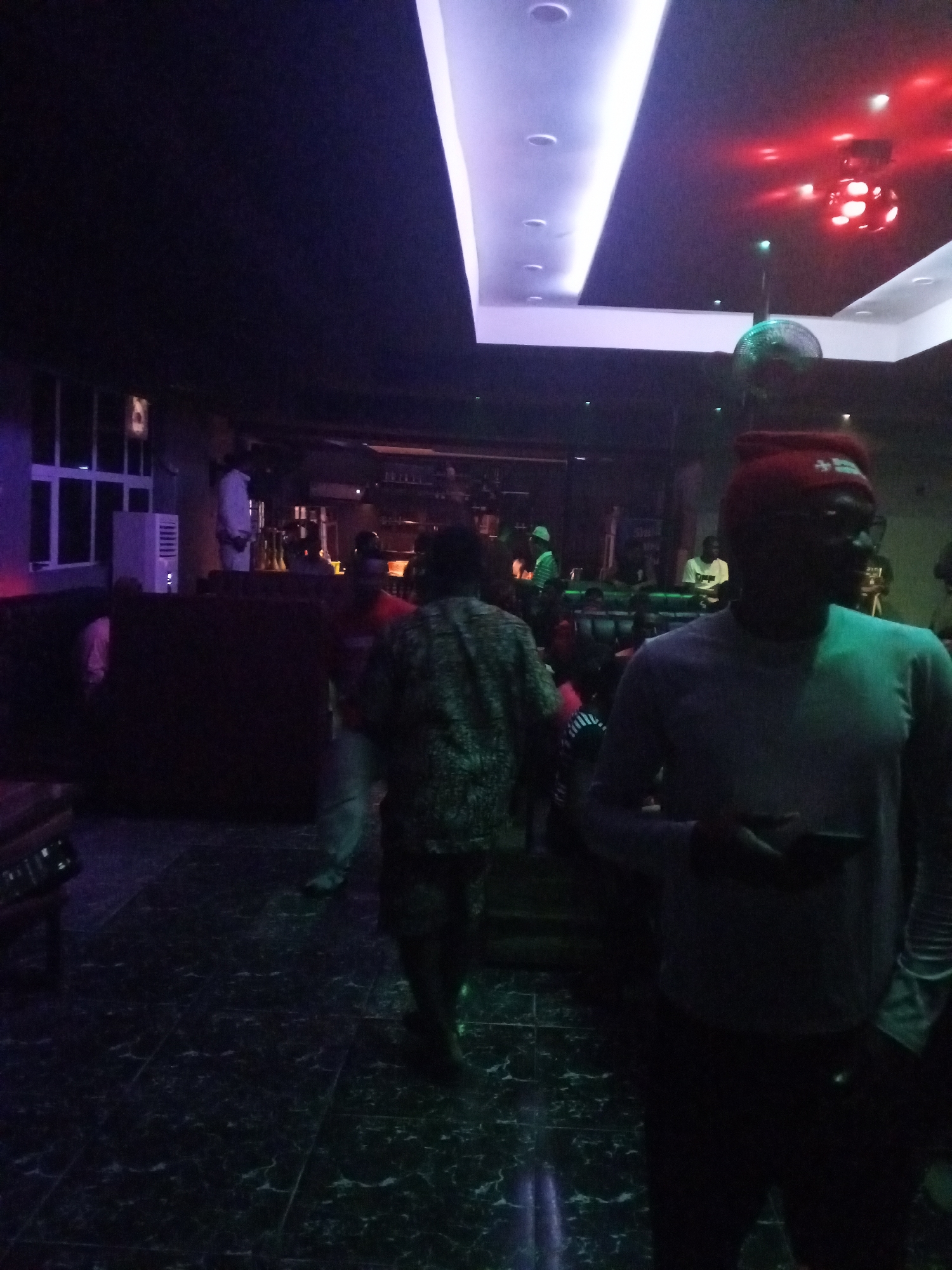 The image depicts a dimly lit interior, possibly a nightclub or lounge, in Unknown, Unknown. The scene features several people; one individual, wearing a red beanie and glasses, stands prominently in the foreground holding a cell phone. Behind him are other patrons, including a man in a patterned shirt. In the background, a bar area is visible, staffed by a person in a white shirt. The lighting is provided by ceiling fixtures including a strip of recessed lights and a disco ball projecting red light. The floor is a dark, patterned tile. There are couches and other seating areas.