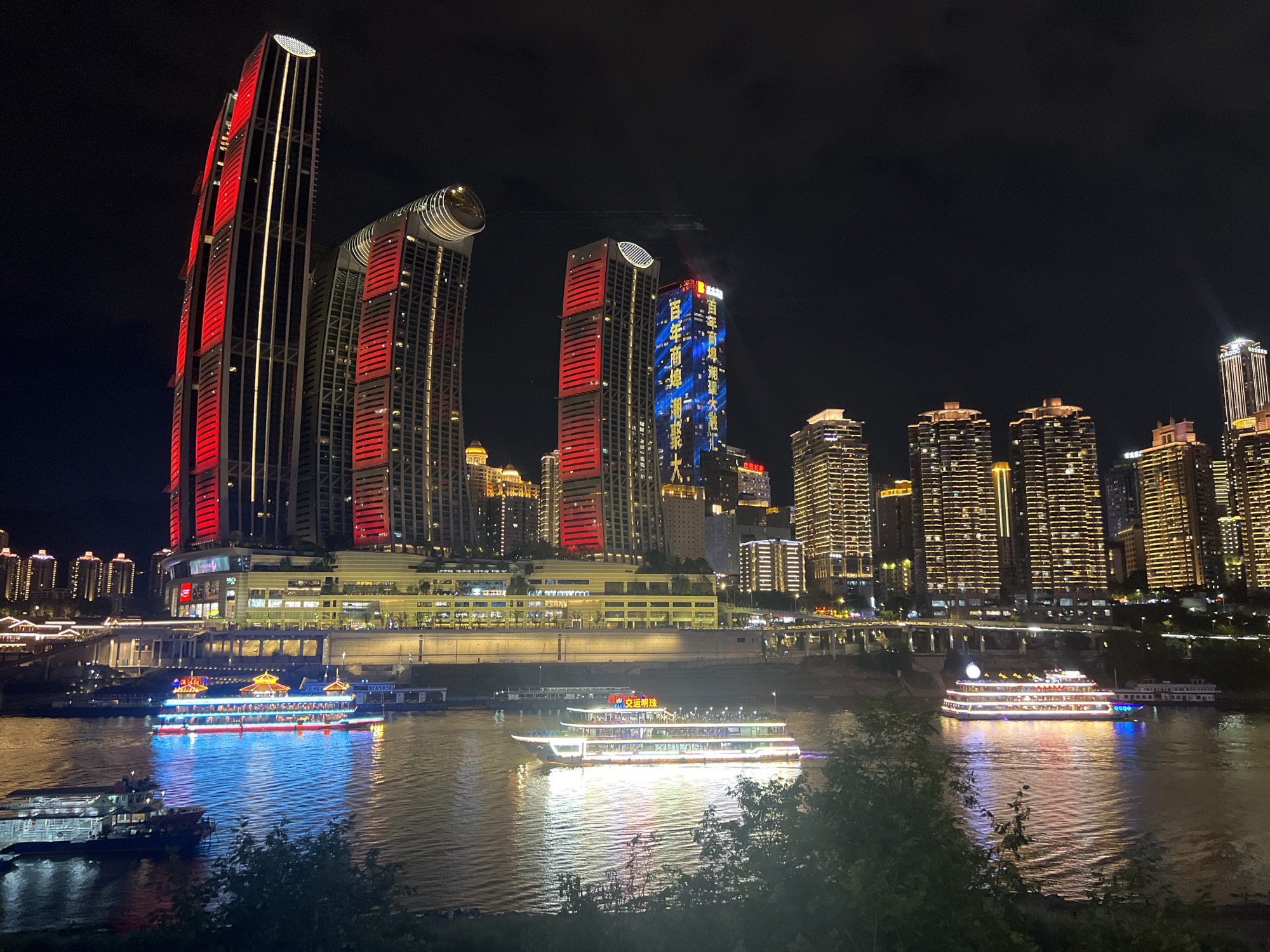 The image captures a nighttime view of the Jiangbeicheng Street area in China, characterized by its illuminated skyline and waterfront. In the foreground, a wide river reflects the vibrant lights of the city and several boats. Two distinct passenger boats are prominently featured on the water. The larger boat in the center is a multi-deck vessel adorned with colorful lights in red, blue, and white, with its reflection shimmering on the water's surface. A smaller, darker boat is positioned to the left, also with visible lights.
The midground showcases a riverside promenade lined with buildings and illuminated structures. Above this, the cityscape is dominated by a collection of towering skyscrapers. Several of these buildings are strikingly lit, with one large skyscraper on the left featuring prominent vertical red lighting, interspersed with white lights. Another skyscraper to its right has a curved, illuminated top section. Further to the right, a skyscraper is adorned with blue and yellow digital displays that appear to form Chinese characters. Numerous other buildings in the background are illuminated with warm, golden light, creating a dense urban landscape. Bridges with illuminated traffic can be seen spanning across the river in the distance. The sky is dark, indicating nighttime, with a few faint clouds or atmospheric phenomena visible. The overall scene conveys a lively and dynamic urban environment at night.