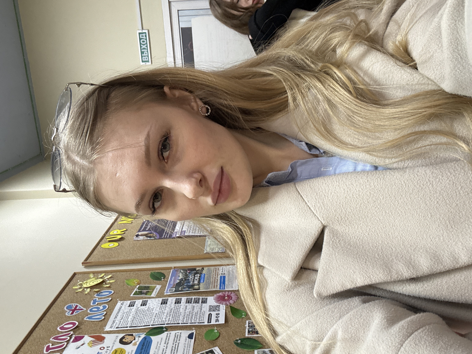 The image is a tilted, close-up portrait of a young woman in an indoor setting, likely an educational institution in Perm, Russia. The woman, who is the primary subject, has long, wavy blonde hair and light-colored eyes, and appears to be wearing light makeup. She is looking directly at the camera with a calm expression. She wears a light-colored (cream or beige) coat over a light blue striped shirt. Sunglasses are perched on top of her head, and a small, circular, embellished earring is visible on her right ear.
In the upper background, another person with dark hair is partially visible. The immediate background consists of a light-colored wall and two cork bulletin boards filled with various papers, flyers, and decorative elements. A green sign with the Russian word "ВЫХОД" (EXIT) is clearly visible on the wall, confirming the Russian-speaking context.
The bulletin boards display several pieces of text and imagery. On the top board, "OUR N" is partially visible. The lower board contains "ЛЕТНИЕ ПРОГРАММЫ" (SUMMER PROGRAMS), "THE LOST WORLD", "АВТО" (AUTO) next to a heart and UK flag, and a phone number "204-42-05". Other fragmented Russian text refers to "профессий" (professions), "лазертаг" (laser tag), "скалодром" (climbing wall), "бассейна" (pool), "экскурсия" (excursion), and "до 31 марта" (until March 31st), along with timings "По будмын 09-30-14:30" (On weekdays 09:30-14:30). These details suggest the boards are used for announcements related to school activities or local events. The overall scene captures a casual moment, possibly a selfie or a close-up photograph taken during a school day.