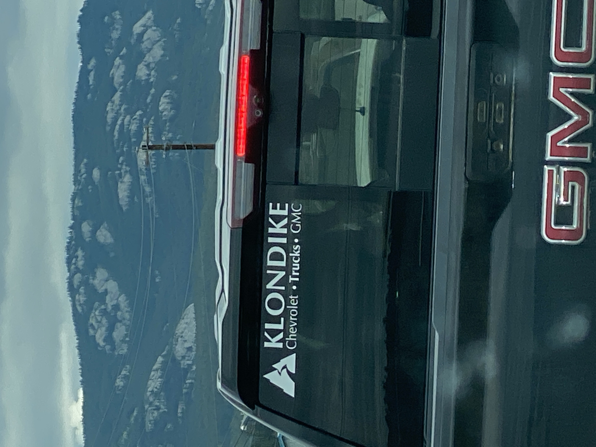 The image captures a rear view of a dark-colored GMC pickup truck, parked in what appears to be an outdoor setting with a mountainous, cloudy backdrop. The truck has a decal on its rear window that reads "KLONDIKE Chevrolet • Trucks • GMC" with a white mountain logo. The truck's third brake light is illuminated red. In the background, a wooden utility pole with wires is visible against a backdrop of rugged, rocky hills and a muted sky. The overall scene suggests a location within or near a natural landscape, possibly on the outskirts of Whitehorse, Canada, given the "Klondike" branding which is strongly associated with the Yukon Territory. There are no people visible in the image.