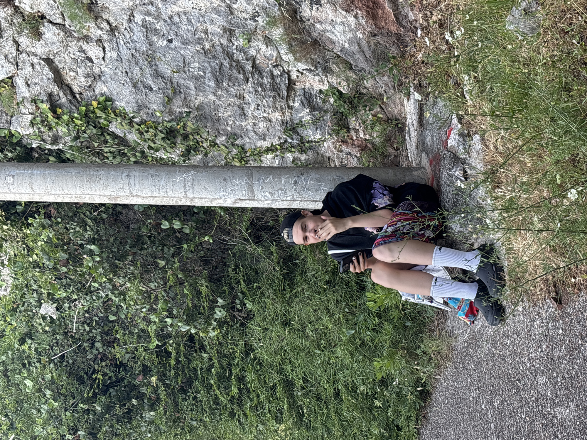 A young person is sitting on a rock next to a concrete pole, holding a phone. They are wearing a black hoodie with purple floral accents on the sleeves, colorful shorts, white socks, and black platform shoes. Their hair is short and they have a black baseball cap on. The background consists of lush green foliage and a rocky cliff face. The ground is covered in gravel and grass. The scene appears to be outdoors, possibly in a natural or park setting.