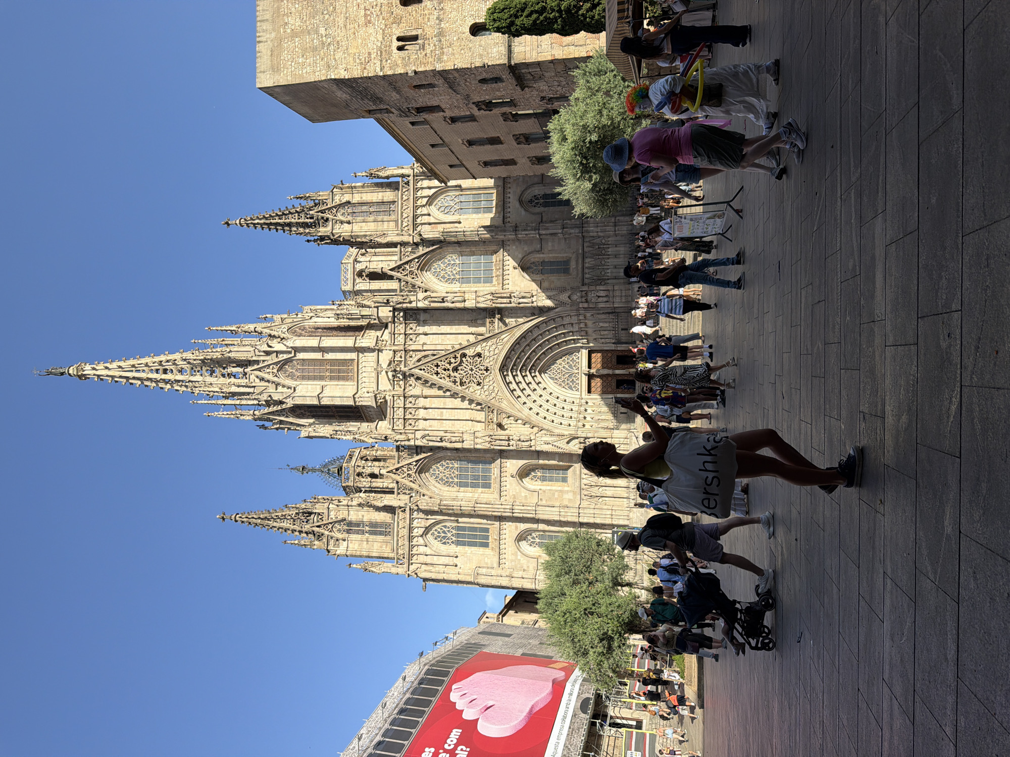 This image depicts a sunny day in Barcelona, Spain, in front of the Barcelona Cathedral. The Gothic cathedral, a prominent landmark, features intricate stonework and soaring spires against a clear blue sky.
In the foreground, a plaza is populated with a diverse group of people. A woman in a yellow top and white shorts is prominently positioned, holding a large white tote bag with "bershka" written on it. She is looking at her phone and appears to be taking a selfie. To her left, a man pushes a stroller with a child inside. A clown with colorful hair, wearing a white costume and holding a balloon animal, is also visible, interacting with another person. Other individuals are scattered throughout the plaza, some walking, some standing, and some appearing to be tourists.
On the left side of the image, a large, red billboard with an image of a hand is partially visible. Behind the people, trees and other buildings, including a section of another stone building to the right of the cathedral, complete the urban scene. The ground is paved with large stone tiles, suggesting a pedestrianized area. The overall atmosphere appears to be one of activity and a typical tourist day in a major European city.