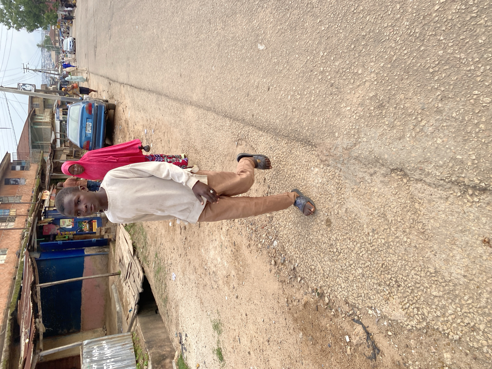 A young boy, appearing to be in his early teens, walks along a dirt and gravel roadside in Jos, Nigeria. He is wearing a long-sleeved, light-colored shirt and brown trousers. On his feet are dark-colored slides. His left hand is extended forward, and he appears to be looking ahead. Behind him, a woman in a vibrant pink hijab walks, her patterned skirt visible below. In the background, a blue sedan is parked on the side of a paved road. Buildings with corrugated metal roofs line the street, and power lines crisscross above. The environment suggests a developing urban or semi-urban area.