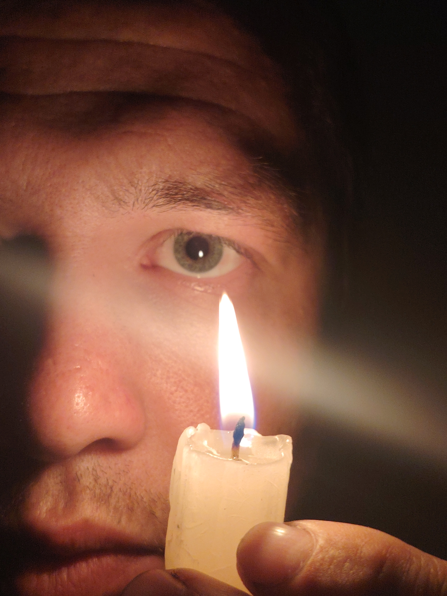 A close-up, low-angle shot captures a person's face in profile, illuminated by the light of a lit candle held in their hand. The focus is on the right eye, which appears to be green or hazel. The skin tone is fair, with some texture and pores visible. The forehead and eyebrows are in the upper portion of the frame, cast in shadow. The nose and mouth are also visible, with a hint of stubble on the chin and upper lip. The candle is a thick, white pillar candle with a slightly uneven top and a visible wick. The flame is bright and casts a warm glow. The background is dark and out of focus. The image does not contain any information about the location in Chaikovsky, Russia.