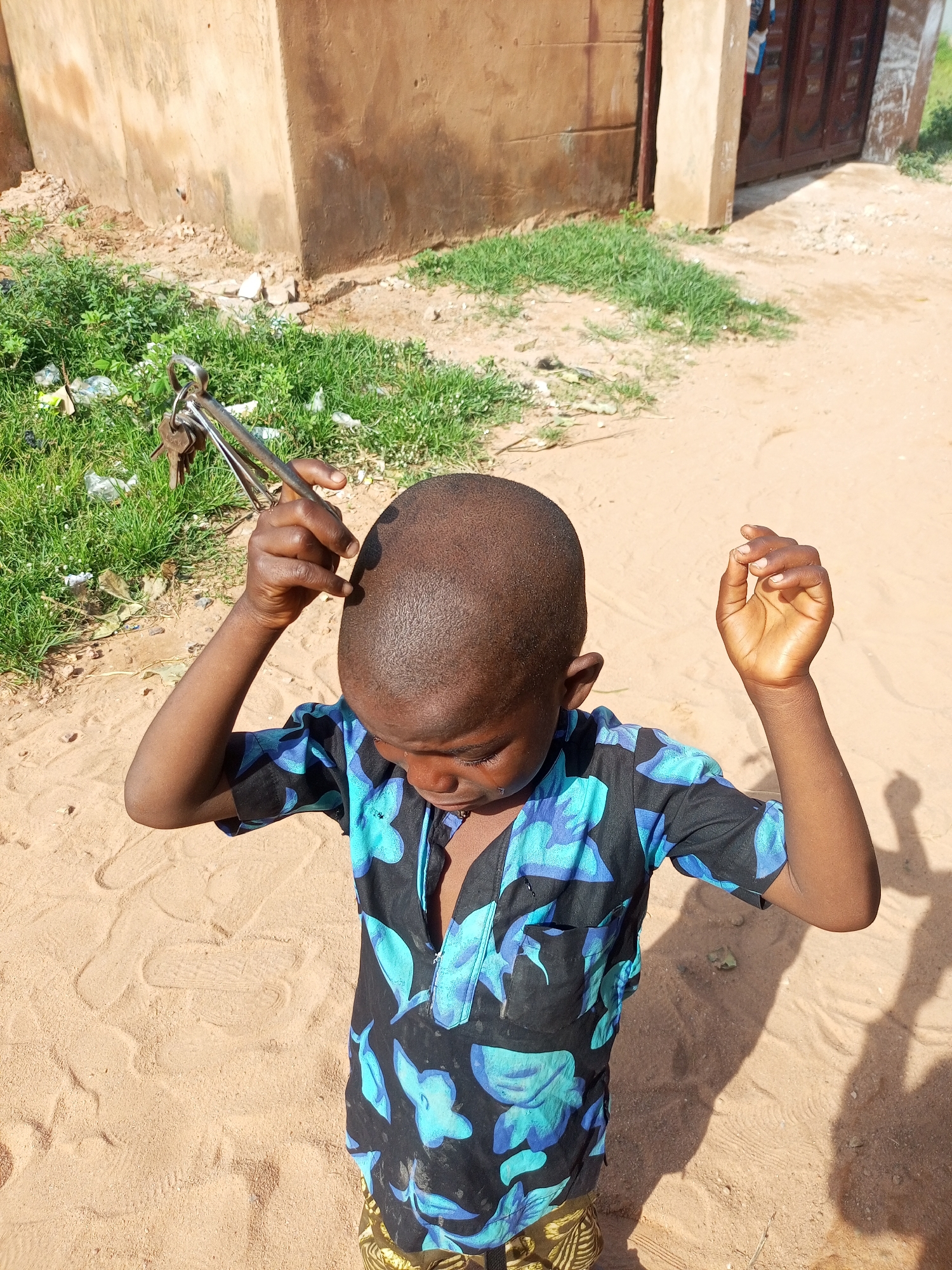 A young boy with a shaved head stands outdoors on a sandy surface, holding a set of keys attached to a metal tool. He is wearing a black shirt with a blue floral pattern and yellow and black patterned shorts. The boy's hands are raised, one holding the keys and the other open. In the background, there is a weathered brown building with a closed metal door and patchy green grass. The scene appears to be in the city of Tempure, Nigeria.