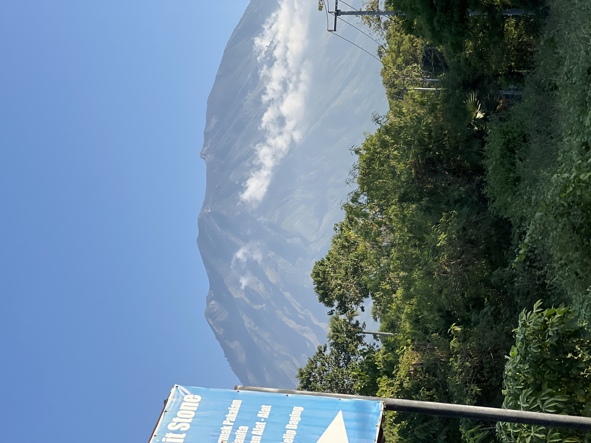 This is a photograph of Mount Agung, an active volcano located in the Karangasem regency in eastern Bali, Indonesia, as seen from Kubu, a village on Bali's northeast coast. The image features the majestic mountain dominating the background, with some clouds clinging to its slopes. In the foreground, there is lush green vegetation, including trees and shrubs, suggesting a tropical environment. To the left, a blue signpost with white text is partially visible, indicating a street-level view and possibly a local business or landmark. The sky above is clear and blue, typical of a sunny day. The overall scene conveys a sense of natural beauty and tranquility, with the imposing volcano serving as a striking focal point.