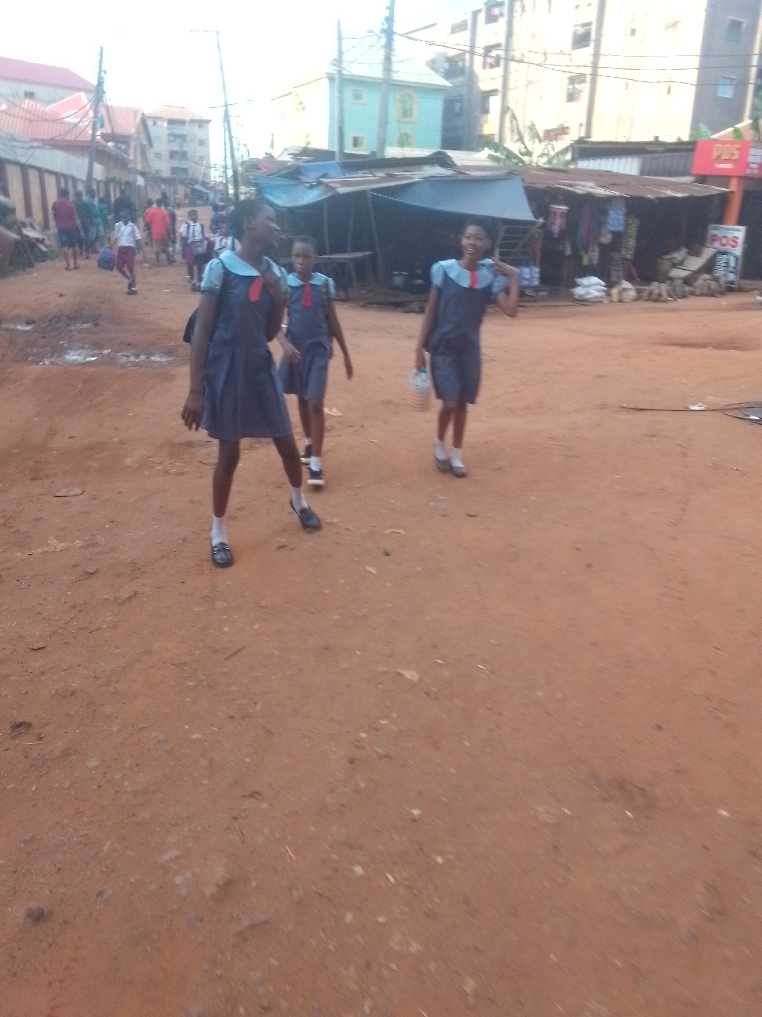 Three schoolgirls in blue uniforms with white socks and black shoes walk along a dirt road in Nkpor, Nigeria. The environment suggests a low-income or developing area, with informal market stalls and simple buildings in the background. Other pedestrians are visible in the distance. A 'POS' (Point Of Sale) sign is visible on one of the stalls. The ground appears dry and dusty.