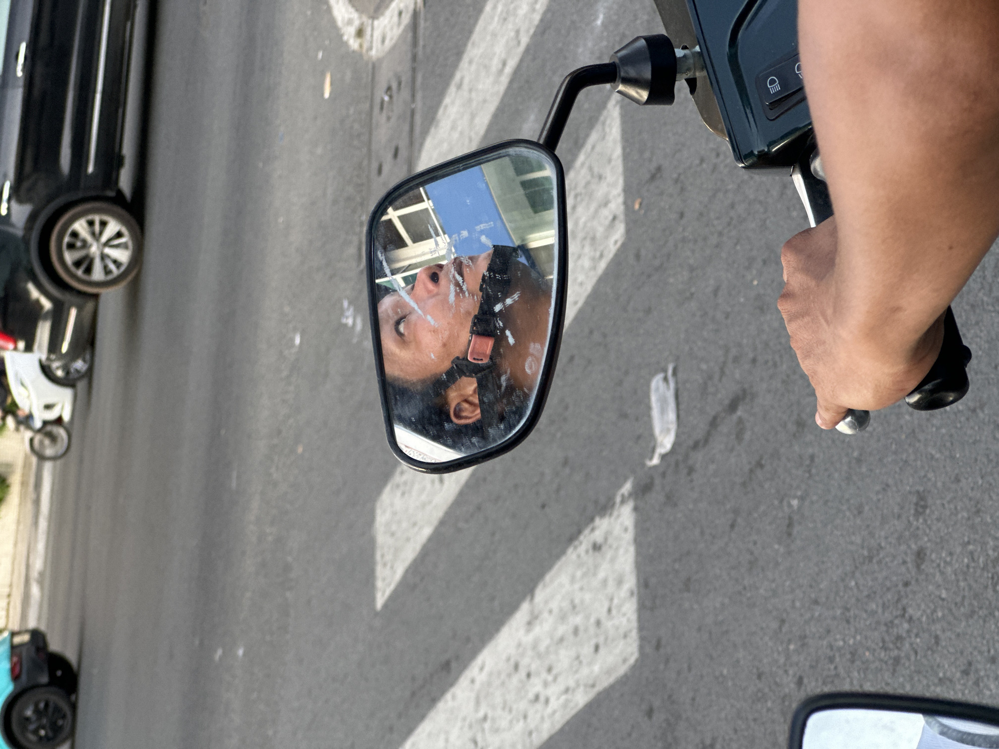 A street in Thủ Đức, Vietnam, seen from the perspective of a person riding a motor scooter. The reflection in the scooter's side mirror reveals the rider's face wearing a face mask and possibly a helmet. A black car and other vehicles, including a light blue scooter, are visible in the periphery. The road has pedestrian crossing stripes. A hand is gripping the scooter's handlebar.