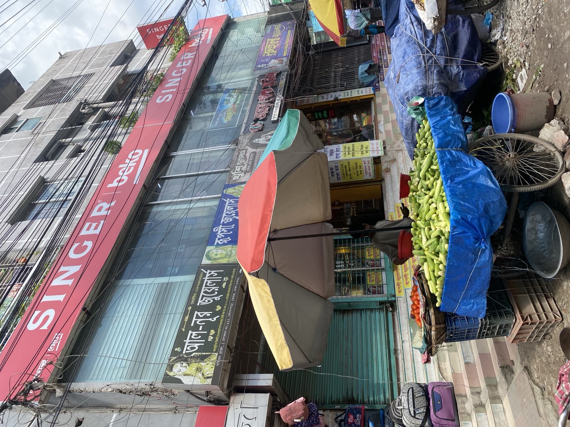 The image captures a street-level view of a commercial building in Dhaka, Bangladesh. A large red awning displays the brand "SINGER" in white lettering, accompanied by the "beko" logo. Below this, the building has large glass windows with roller shutters, some of which are partially open, revealing the interior. Several smaller signs for jewelry shops, such as "আল-নূর জুয়েলার্স" and "রূপসী জুয়েলার্স," are also visible on the facade. In the foreground, a street vendor is selling produce from a cart. A large quantity of cucumbers is displayed on a blue tarp-covered cart, with a basket of tomatoes placed nearby. Two large umbrellas, one red and yellow and the other red and green, provide shade over the vendor's stall. Stacks of luggage and shopping bags are visible on a set of stairs to the left. The street is paved with gravel and dirt, and electrical wires are prominently strung across the building's facade. A man, presumably the vendor, is partially visible behind the produce. The sky appears overcast.