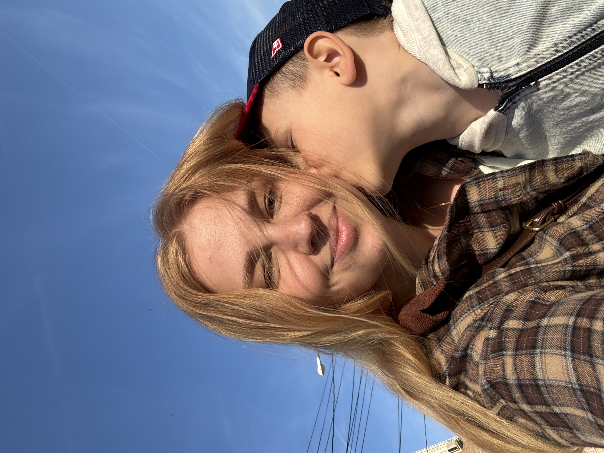 An outdoor selfie, rotated counter-clockwise, captures an intimate moment between a woman and a child under a clear blue sky. A young woman with long, blonde hair and a fair complexion occupies the right side of the frame, looking towards the camera with a soft smile. She is dressed in a brown and tan plaid flannel shirt. Behind and above her, a child, likely a boy, leans in with his face close to her cheek and ear, suggesting an affectionate kiss or whisper. The child wears a dark blue baseball cap with a red brim and a white logo, paired with a light blue denim jacket over a lighter inner shirt.
The background consists primarily of a bright, cloudless blue sky, indicative of sunny weather. Faint white contrails are visible in the upper left portion of the sky. Strong sunlight illuminates the subjects, highlighting their hair and skin. A small, white, possibly plastic earbud or similar object hangs from the woman's hair near her left ear. Thin, dark lines, resembling wires or ropes, are faintly visible against the sky in the lower part of the image. The very bottom right edge of the frame shows a slight, indistinct outline of a white building structure. The overall mood of the image is tender and joyful, reflecting a strong bond between the two individuals.